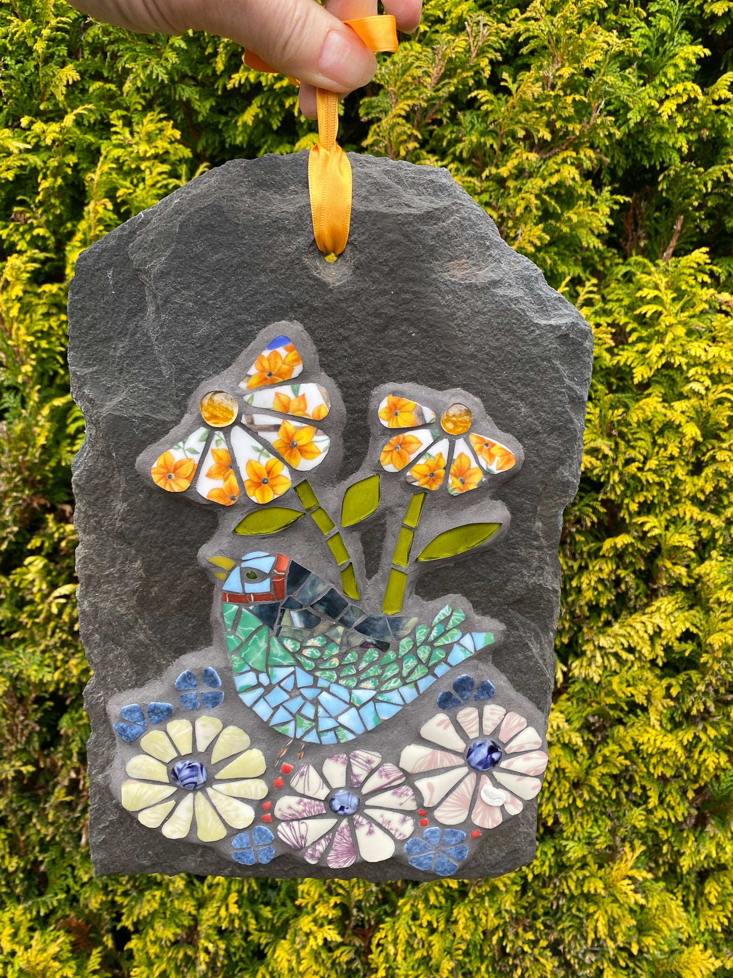 mosaic on slate with a green and blue bird, with purple and yellow flowers.  The petals are patterned from the crockery used.  Photographed with a foliage background