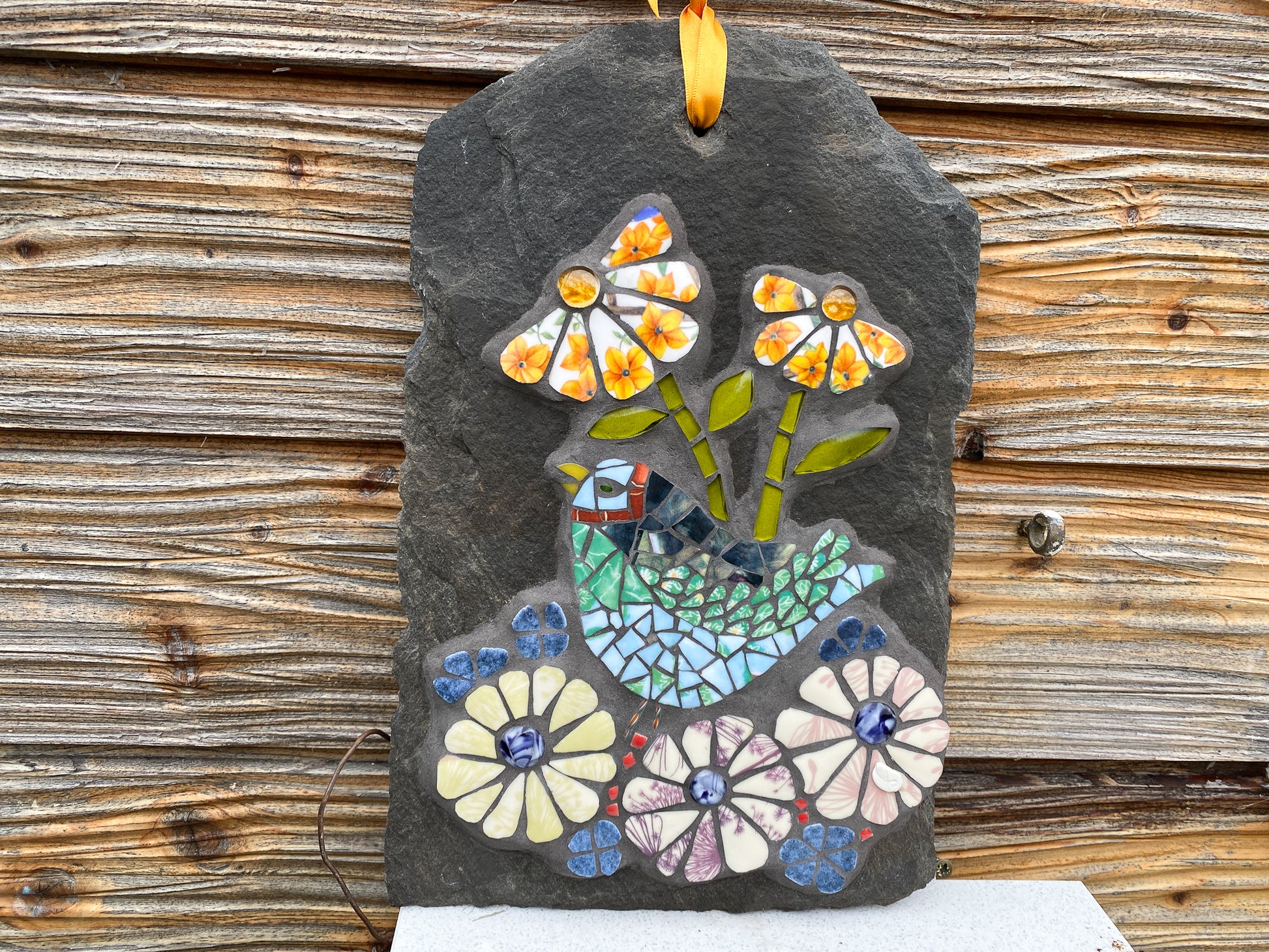 mosaic on slate with a green and blue bird, with purple and yellow flowers.  The petals are patterned from the crockery used.  Photographed with a wooden shed background