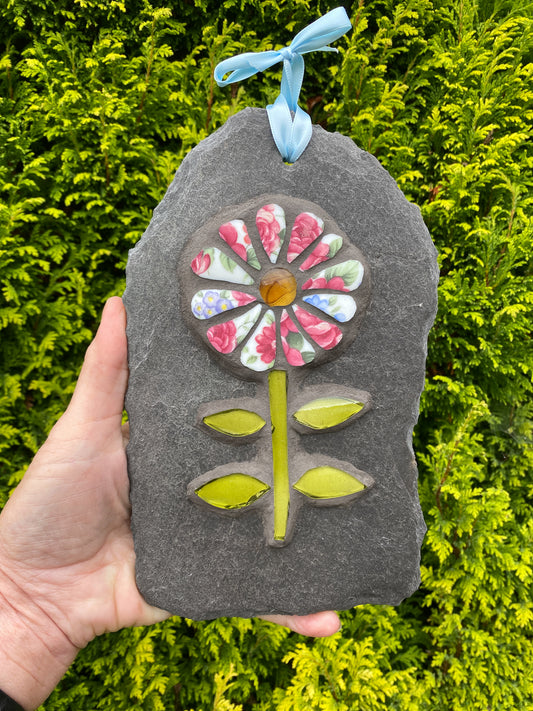 A hand holds a dark gray stone garden ornament shaped like a flower, decorated with colorful floral-patterned petals, a yellow center, green stem and leaves, and a light blue ribbon loop at the top, set against a background of green foliage.