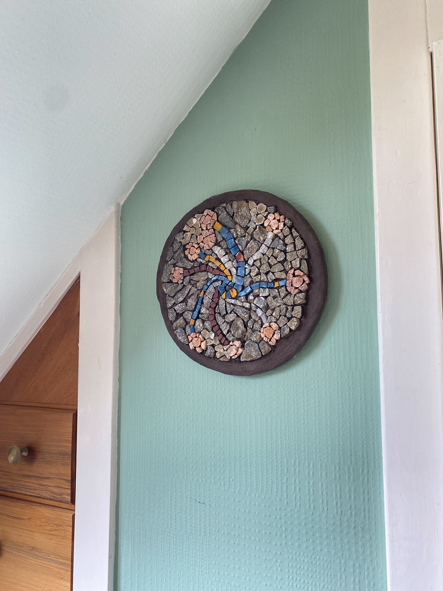 A circular mosaic made of various stones, with threads of blue and yellow plastic, and quartz representing stems, and pink toned stones for flower heads.  Brown painted edging.  Shown on a soft green internal wall 