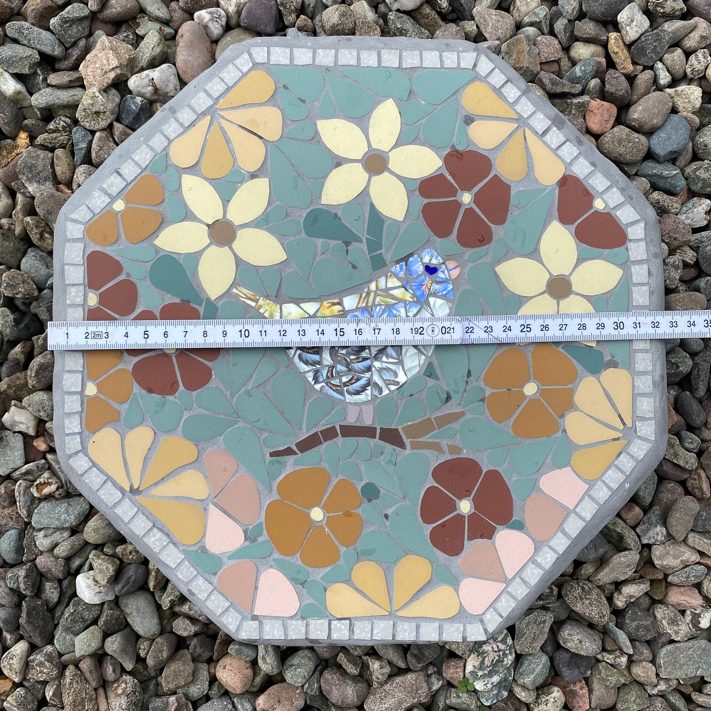 An octagonal mosaic garden stone with a floral pattern and a small bird motif, shown on a bed of gray pebbles with a tape measure across the middle to indicate size.