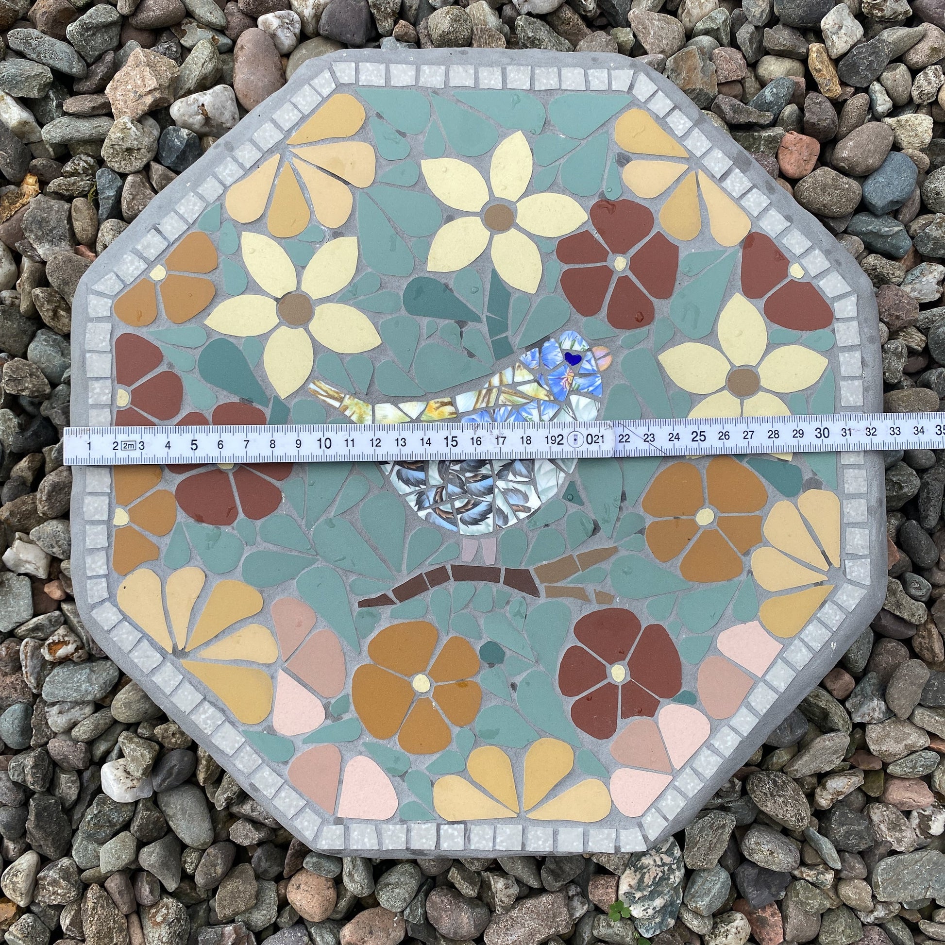 An octagonal mosaic garden stone with a floral pattern and a small bird motif, shown on a bed of gray pebbles with a tape measure across the middle to indicate size.