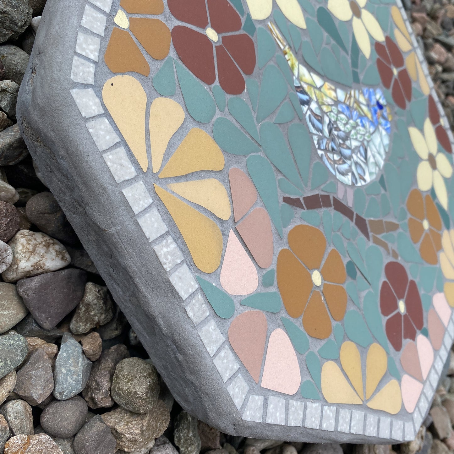 octagonal mosaic garden stone with a bird and floral design, shown resting on a bed of small gray pebbles.