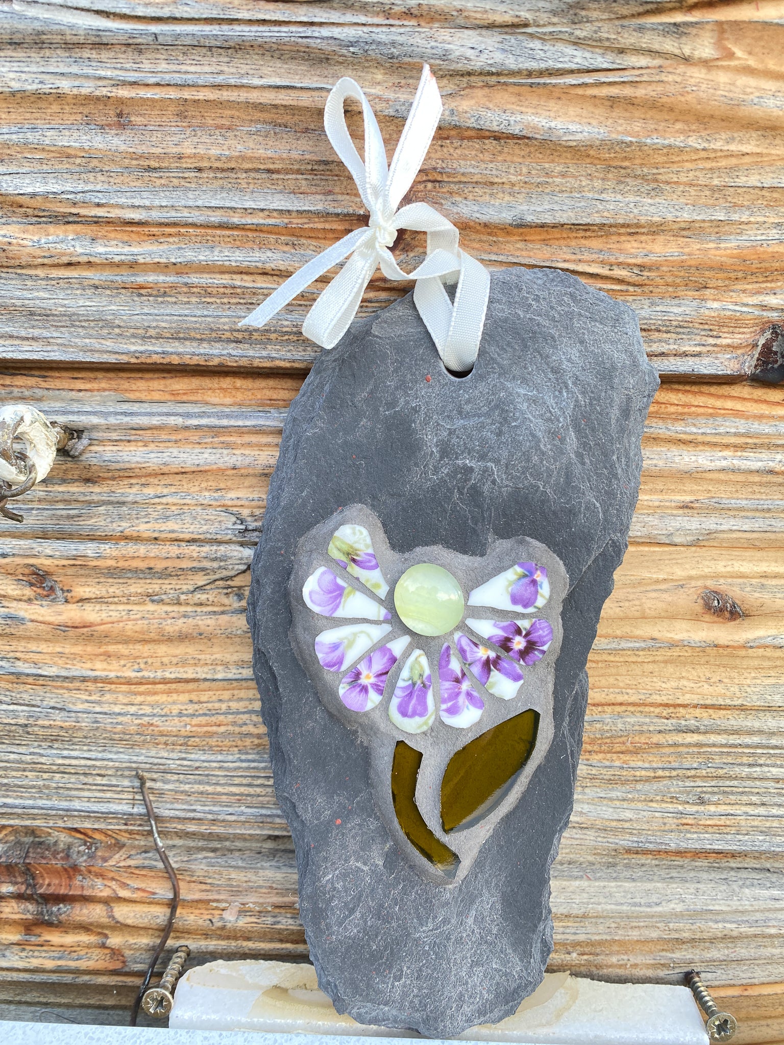 small daisy in mosaic with violet flowers on the petals.  a pale yellow glass nugget at the centre of the flower, dark green stem and leaf. on a pale grey thin slate, shown against a wooden backdrop