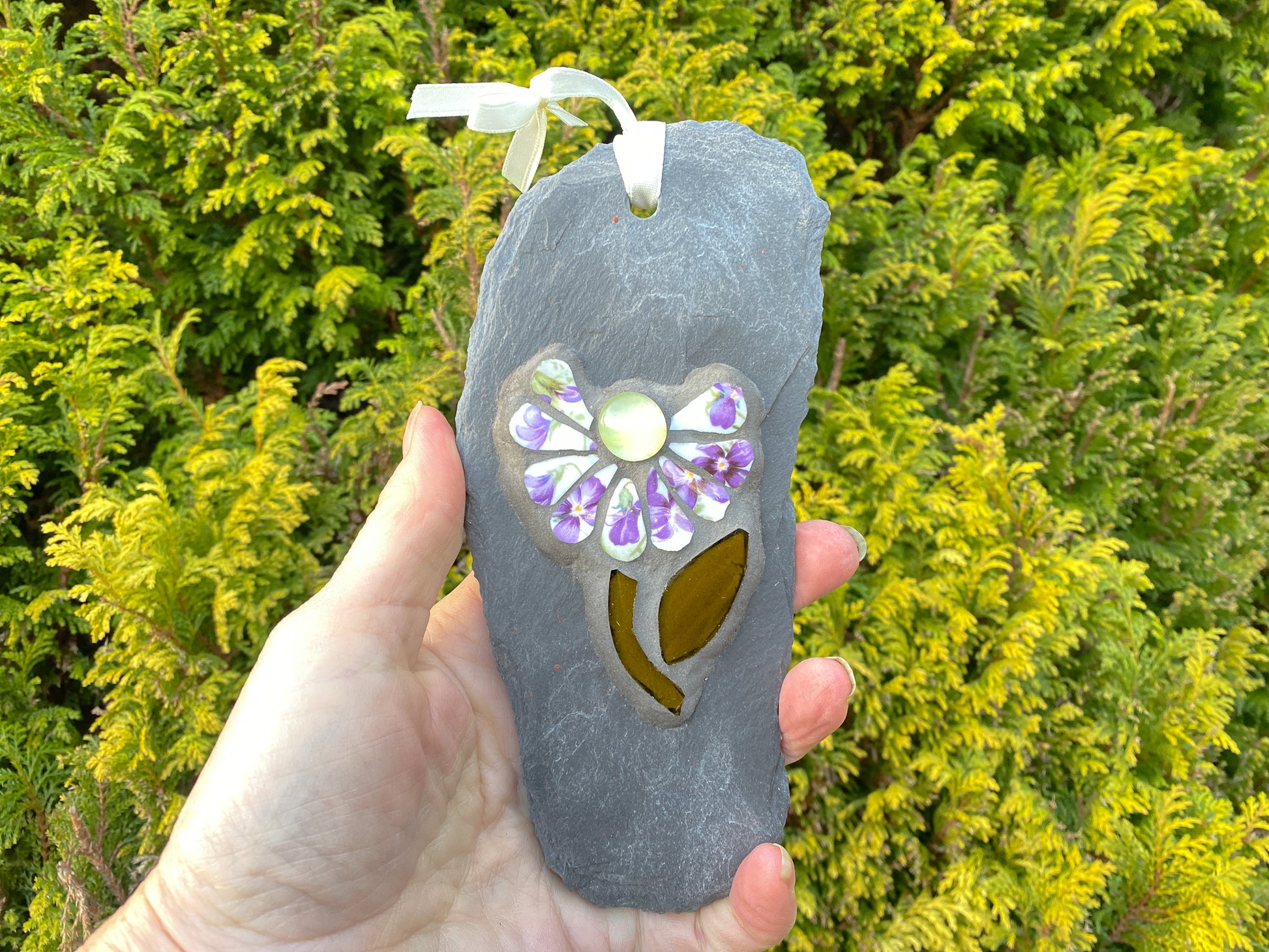 small daisy in mosaic with violet flowers on the petals.  a pale yellow glass nugget at the centre of the flower, dark green stem and leaf. on a pale grey thin slate, held in the hand