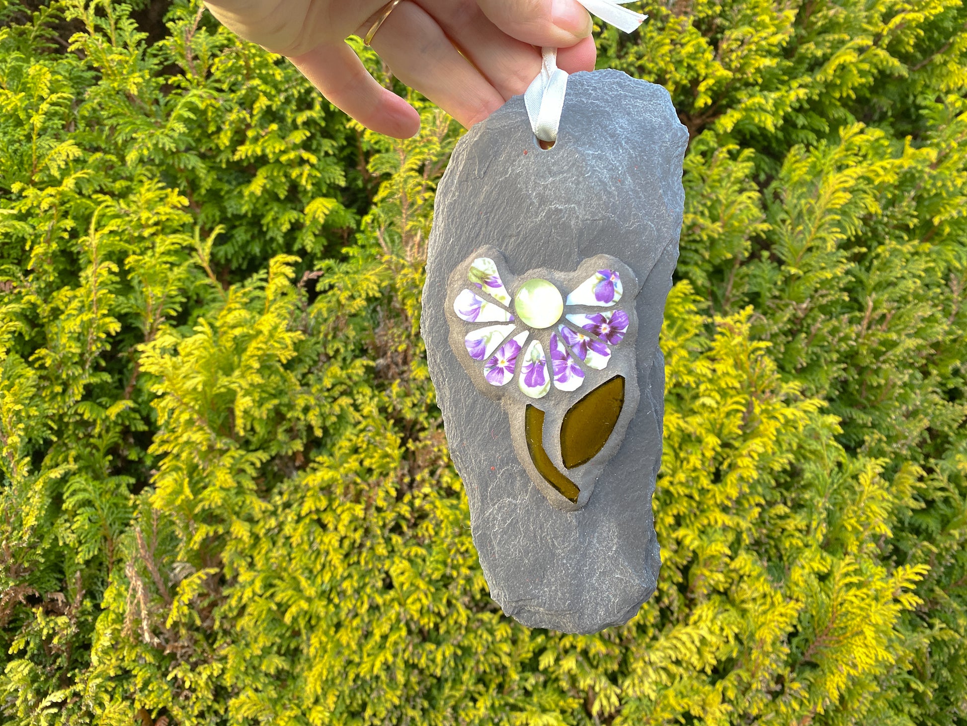 small daisy in mosaic with violet flowers on the petals.  a pale yellow glass nugget at the centre of the flower, dark green stem and leaf. on a pale grey thin slate, held by a ribbon from above