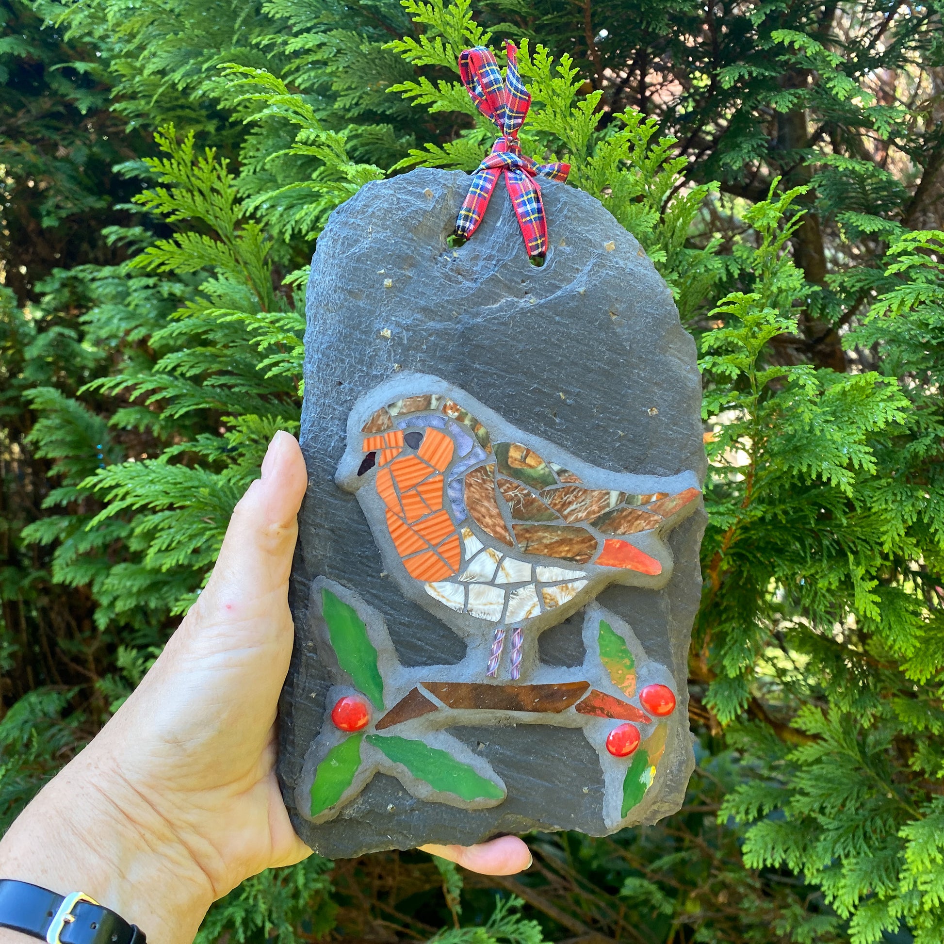 Decorative slate with mosaic design of a robin held by a hand against a green foliage background