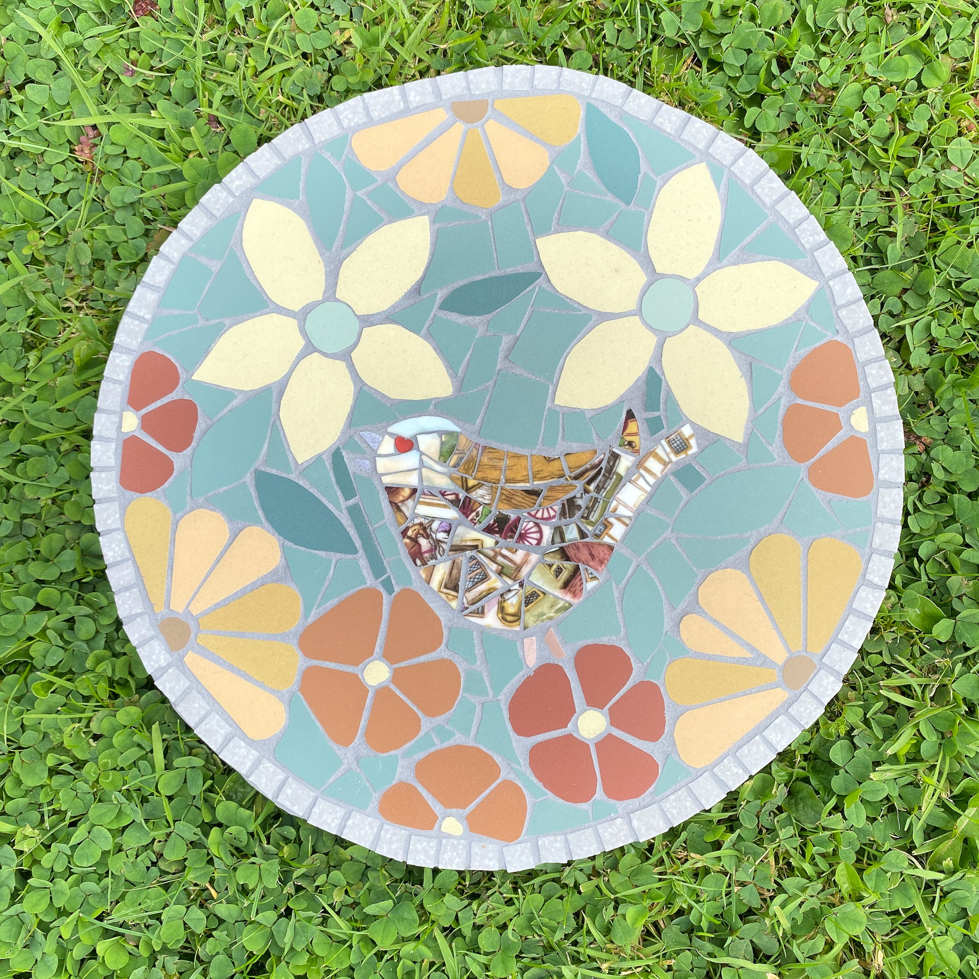 A round mosaic garden stone featuring a bird silhouette in the center with a reflective silver mirror-like surface, surrounded by a floral pattern in light blue, yellow, orange, and red on a green grass background.
