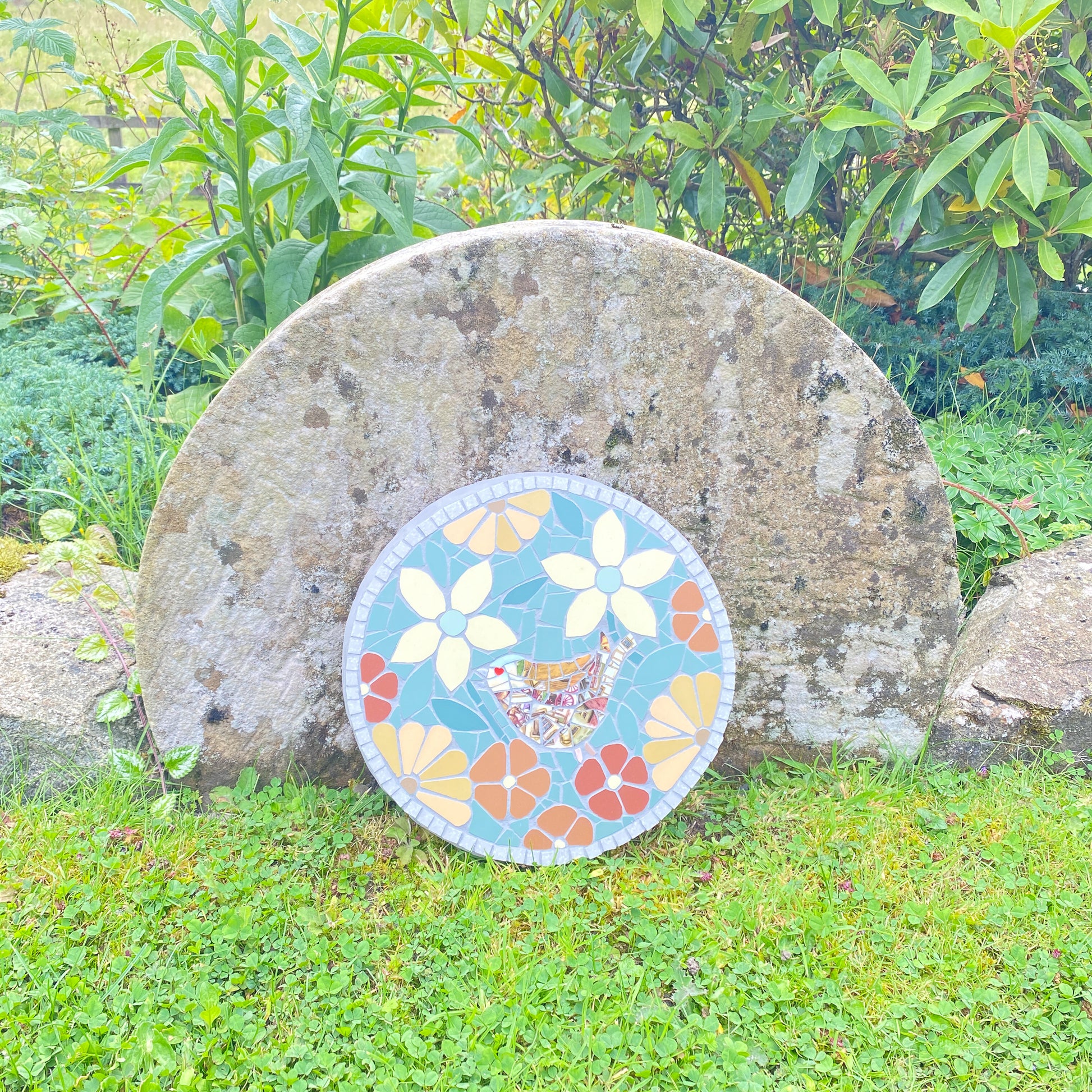 A round mosaic garden plaque featuring a colorful bird and floral design, displayed outdoors against a large natural stone on a grassy lawn with green foliage in the background.