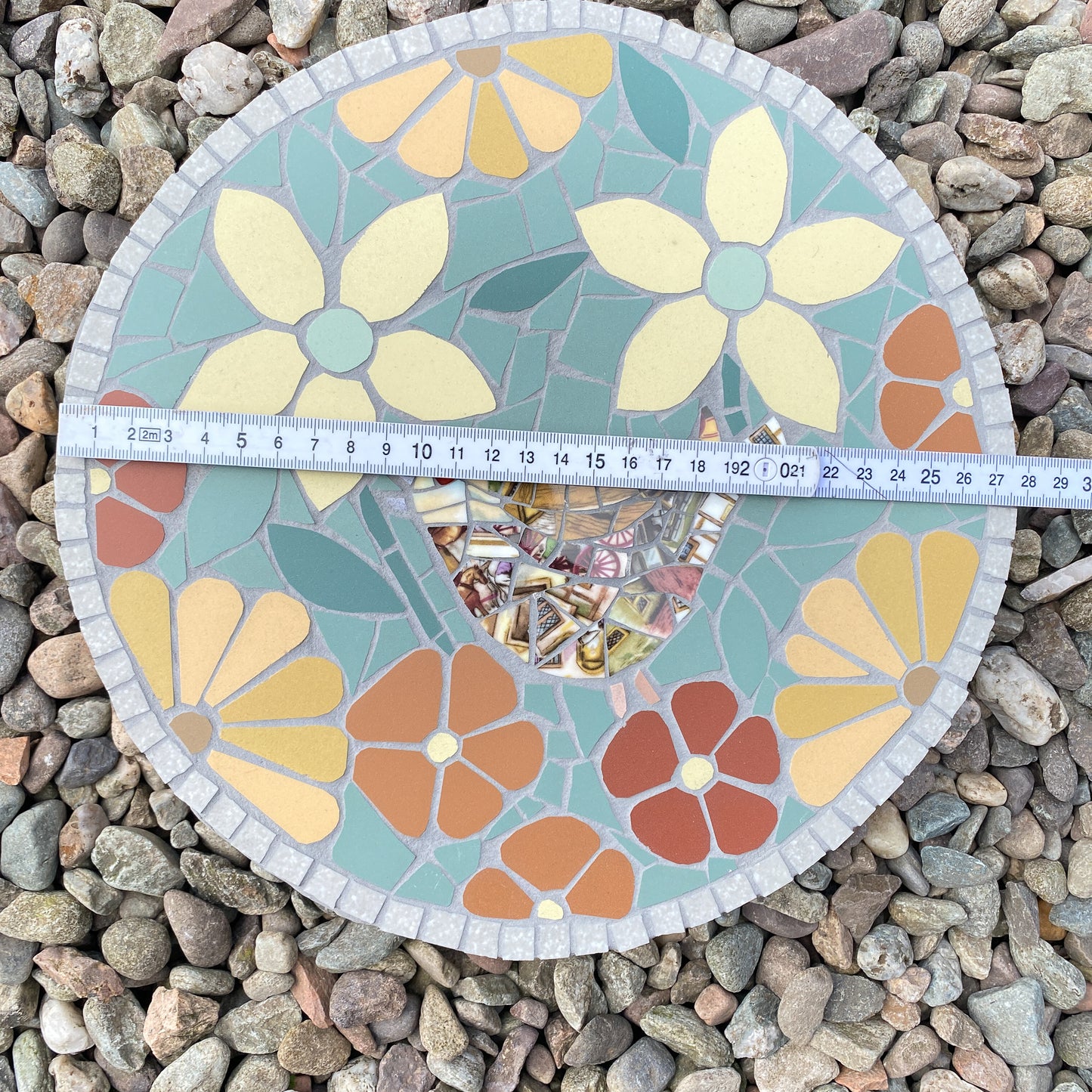 A round mosaic garden stone featuring stylized flower designs in yellow, orange, and red on a light blue-green background, shown on a bed of small gray pebbles with a measuring tape across the middle to indicate size.