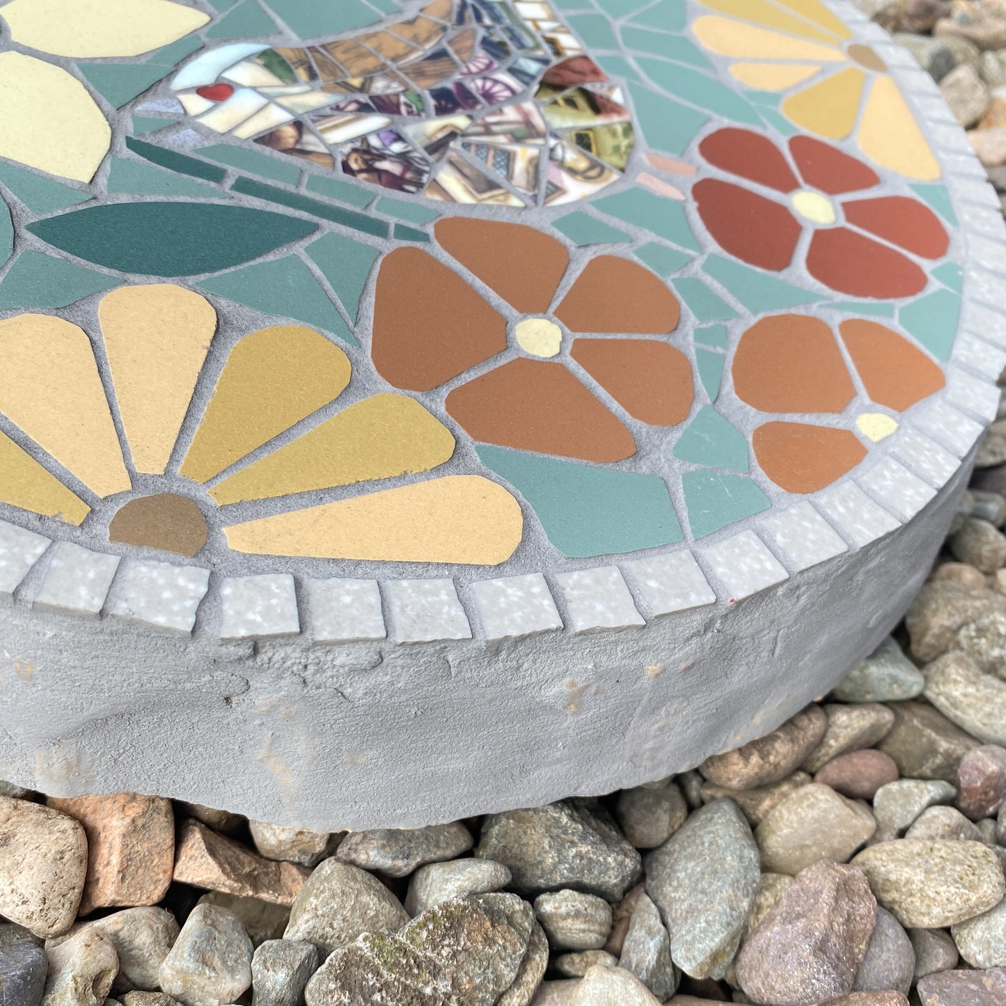 A round mosaic garden stone with a floral pattern, placed on a bed of small gray and brown pebbles. The stone features teal, yellow, orange, and brown tiles with a gray concrete base.