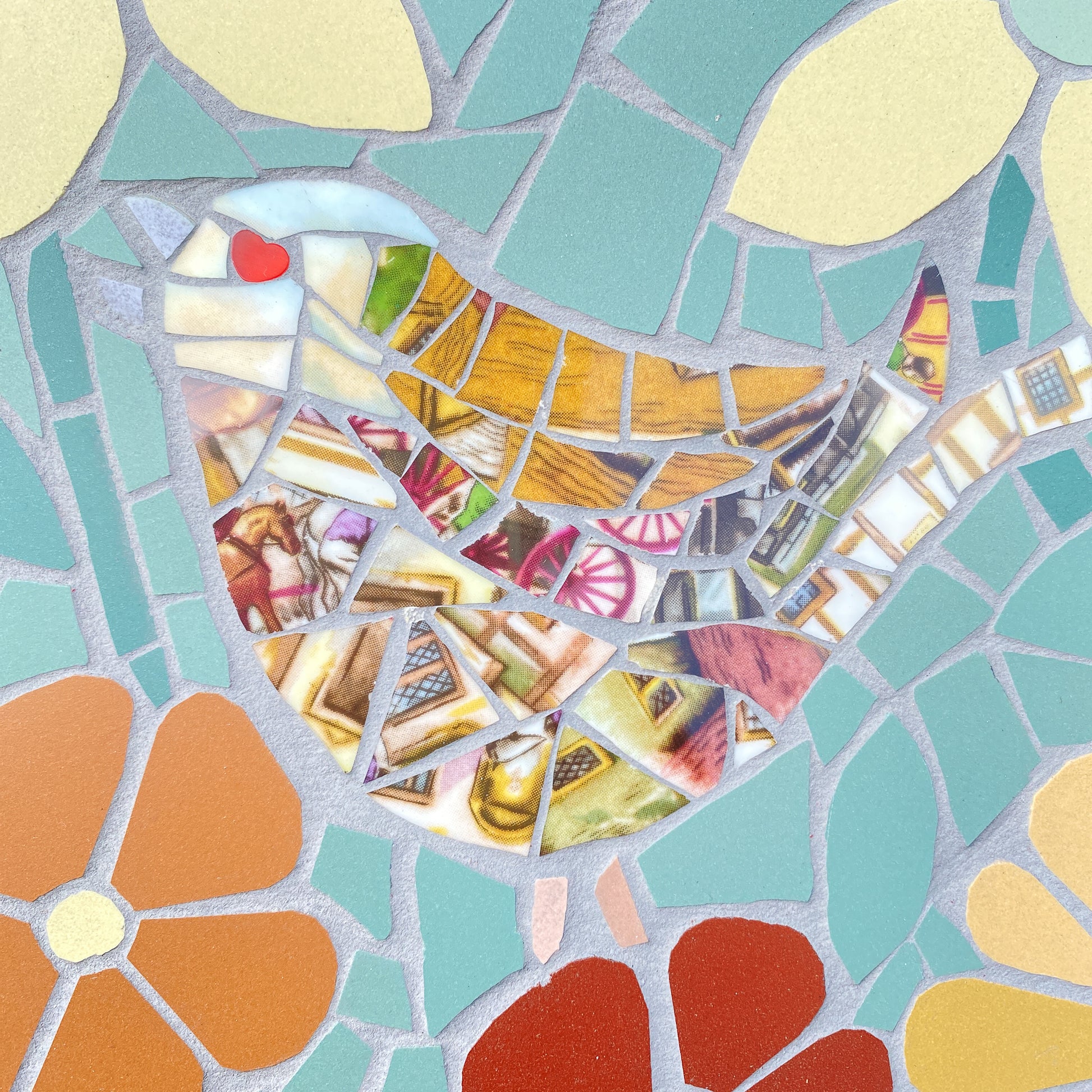 A colorful mosaic bird artwork made from many small cut pieces in turquoise, cream, orange, red, yellow, pink, and brown, with a gray stone-like grout outline.
