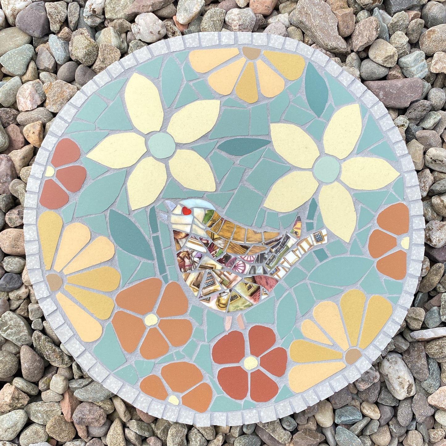A round mosaic garden stone featuring a bird and floral design, placed on a bed of small gray river rocks. The mosaic is made of pastel blue, yellow, orange, and red pieces with a gray border.