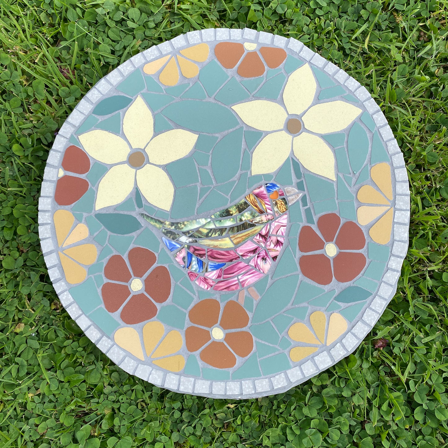 A round mosaic garden stone featuring a colorful bird and floral design, placed on green grass.