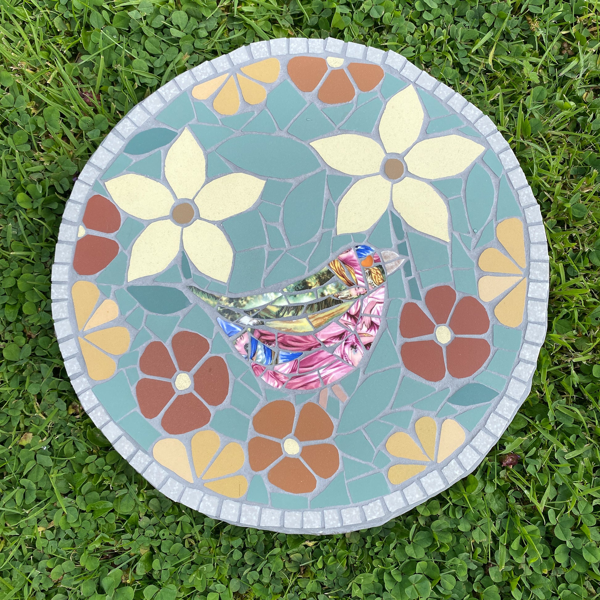 A round mosaic garden stone featuring a colorful bird and floral design, placed on green grass.