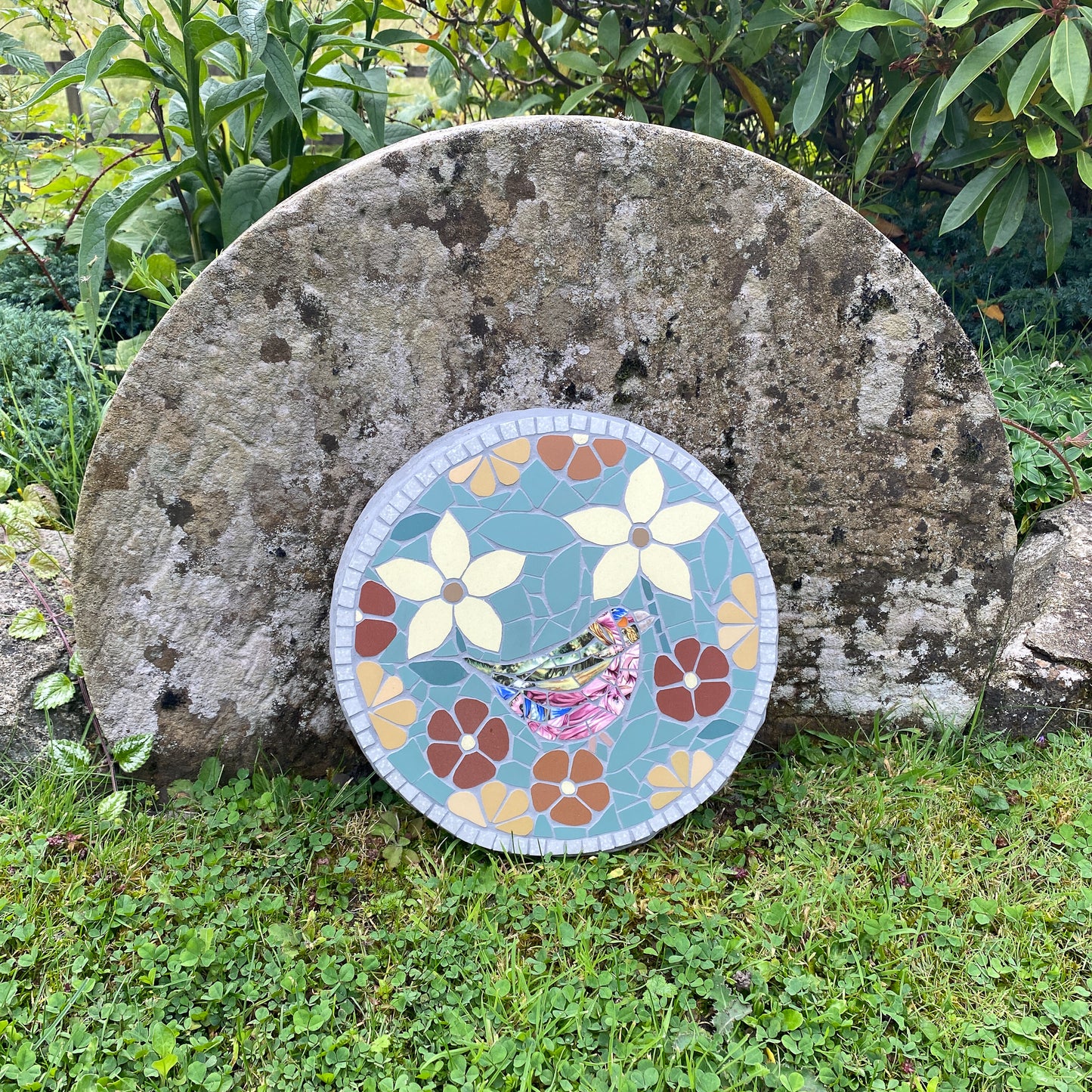 A round mosaic garden stone plaque featuring a bird and floral pattern, leaning against a large natural stone in a grassy outdoor garden setting.