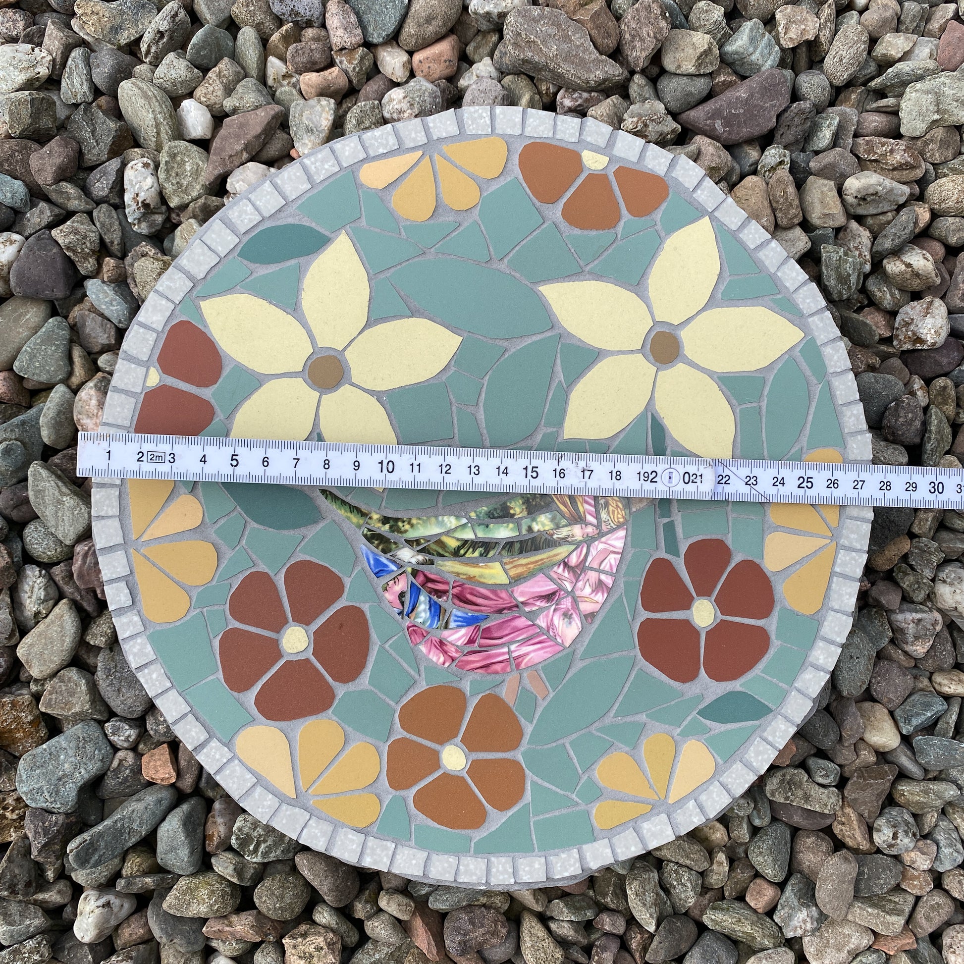 A round mosaic tile plaque featuring a stylized bird and floral motifs, laid flat on a bed of small gray stones with a measuring tape across the top to show dimensions.