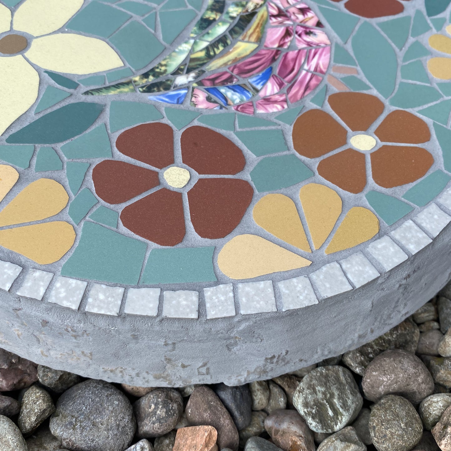 A close-up of a round mosaic garden stone or planter base with colorful flower tiles, set on a bed of small gray stones. The mosaic features teal, brown, yellow, pink, and iridescent green-blue tiles.