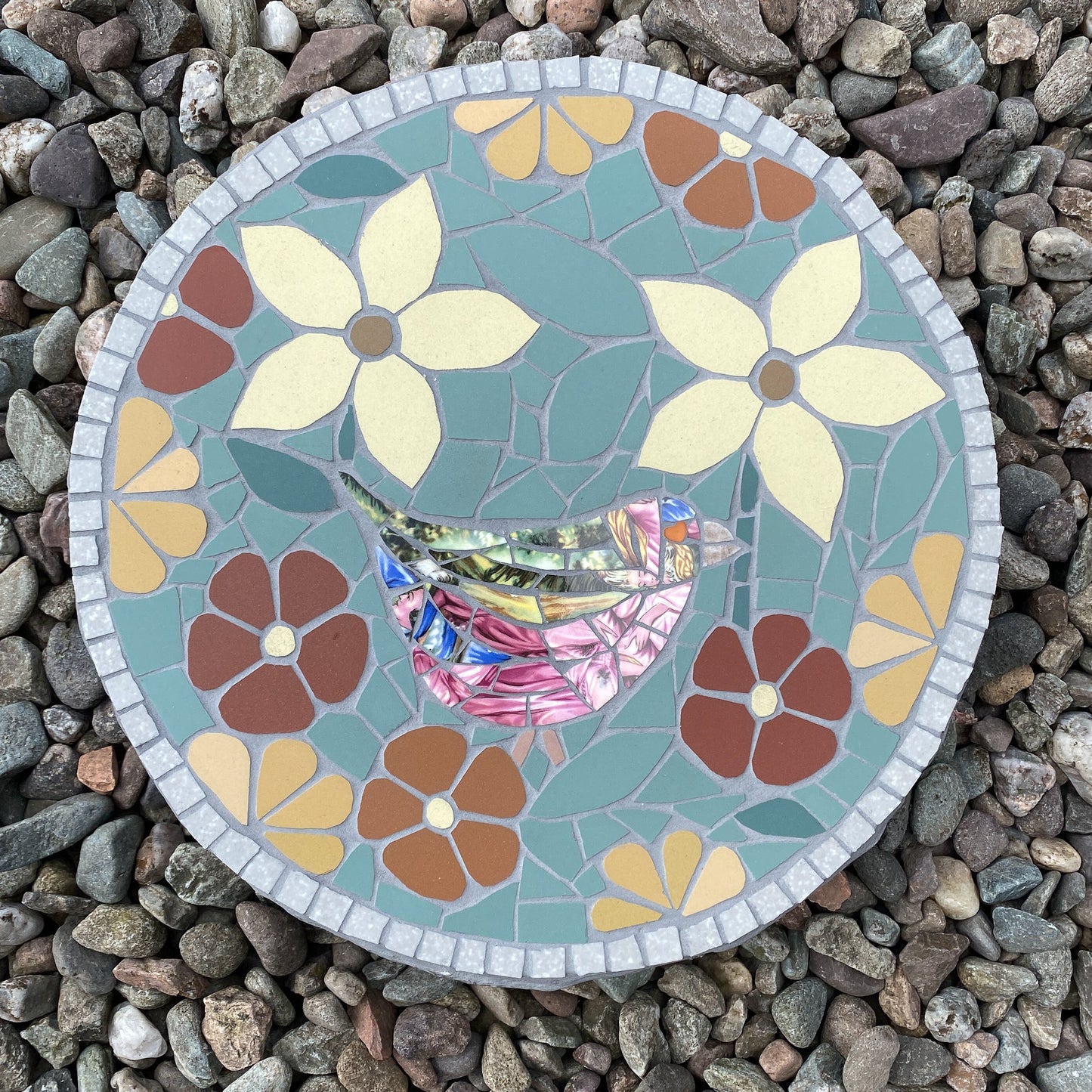 A round mosaic garden stone featuring a colorful bird and floral design, placed on a bed of small gray pebbles.