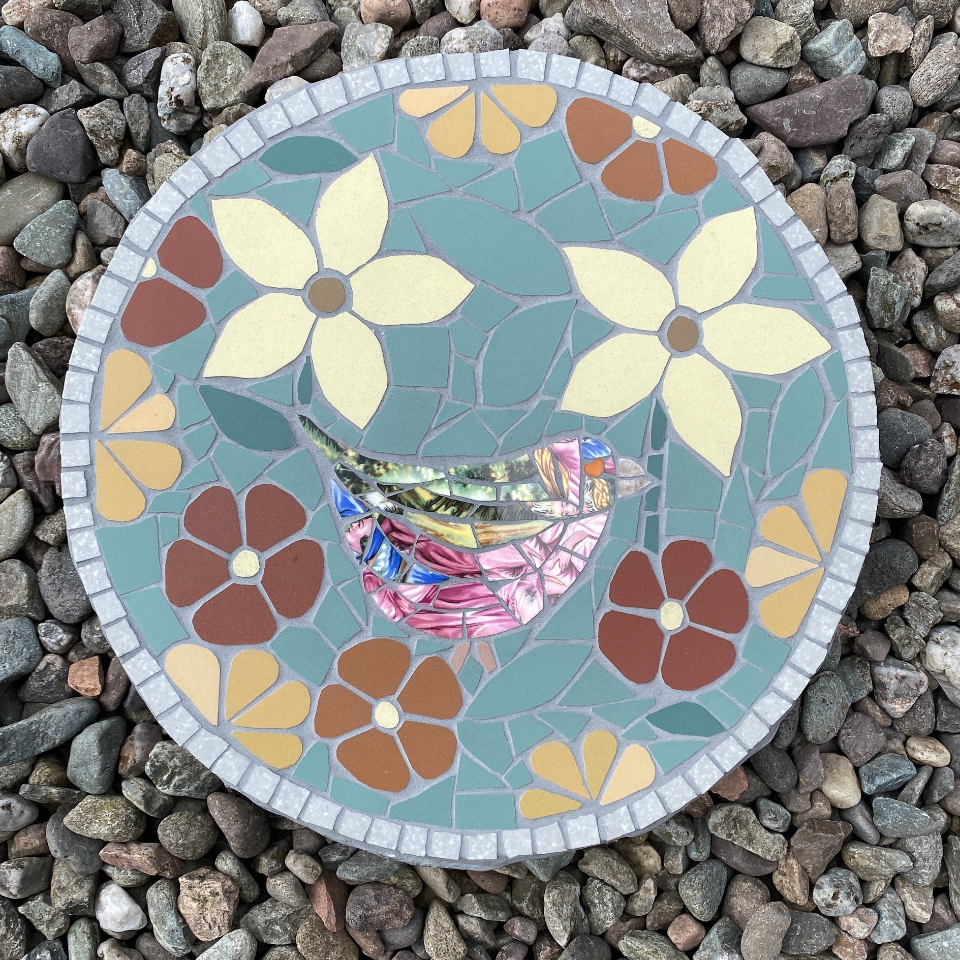 A round mosaic garden stone featuring a colorful bird and floral design, placed on a bed of small gray pebbles.