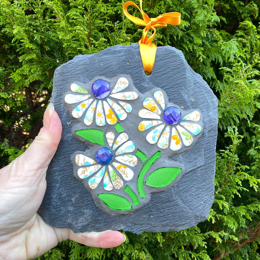 three mosaic flowers with purple blue glass nuggets at the centre.  petals are in tangerine, blue and white coloured, with stem bright green, on a grey slate. shown hand held against an evergreen tree