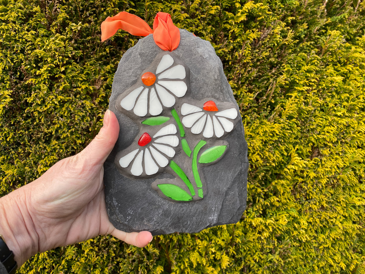 Three white daisies in mosaic on a grey slate.  Each daisy has seven petals and a vibrant orange nugget. bright green stem and leaves. shown hand held against an evergreen background
