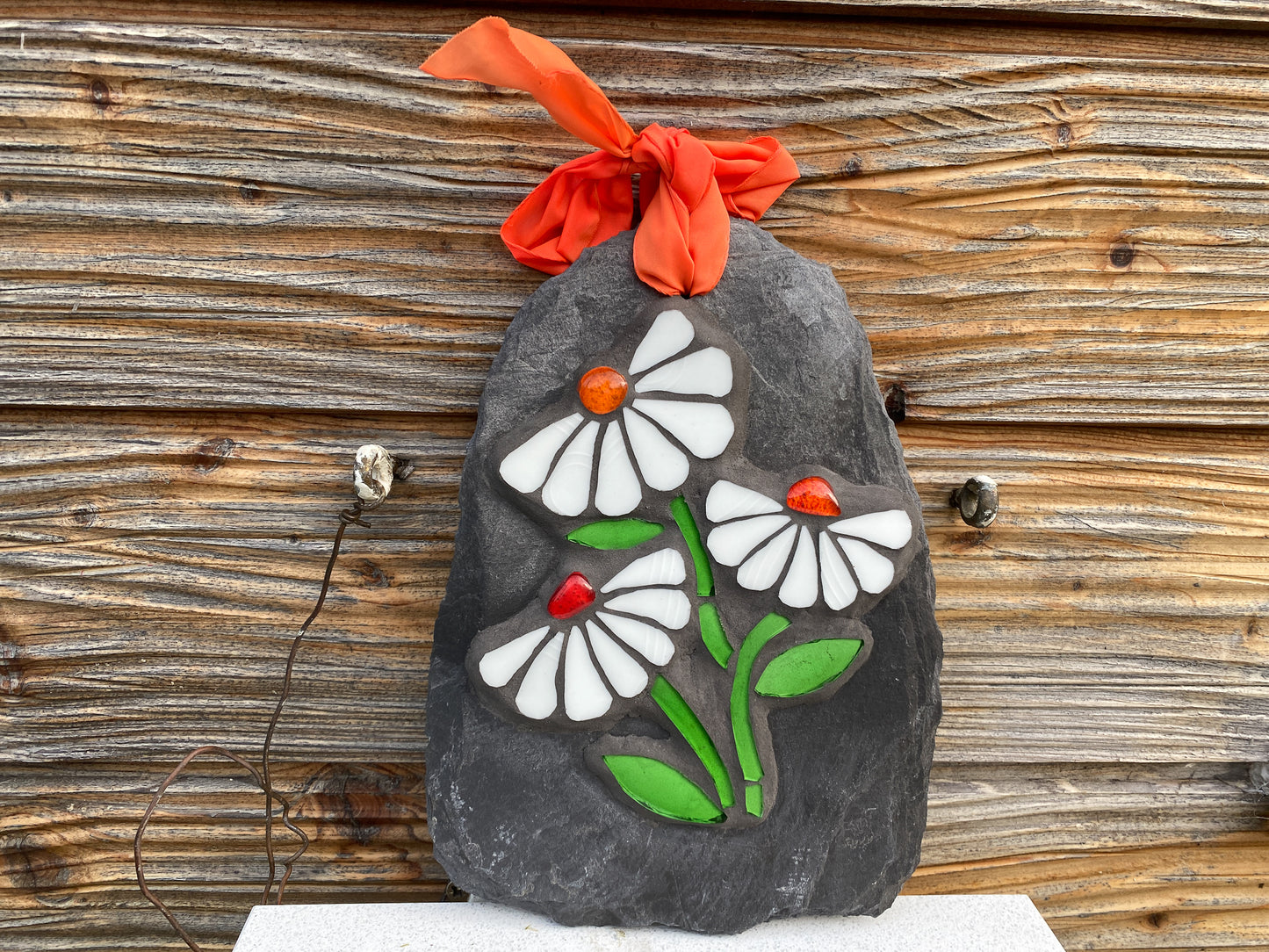 Three white daisies in mosaic on a grey slate.  Each daisy has seven petals and a vibrant orange nugget. bright green stem and leaves. shown against wooden panelling