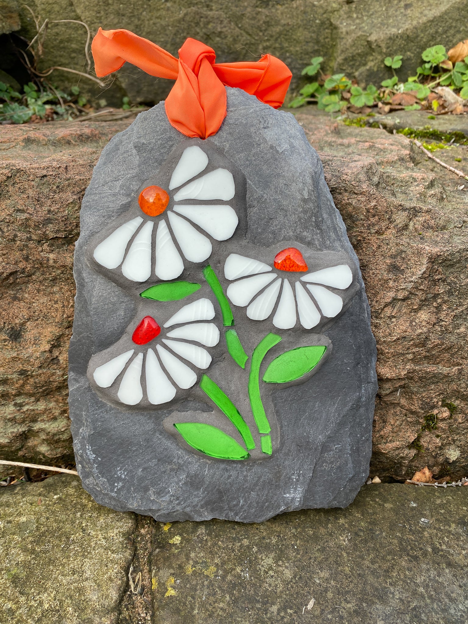 Three white daisies in mosaic on a grey slate.  Each daisy has seven petals and a vibrant orange nugget. bright green stem and leaves. shown on a stone step