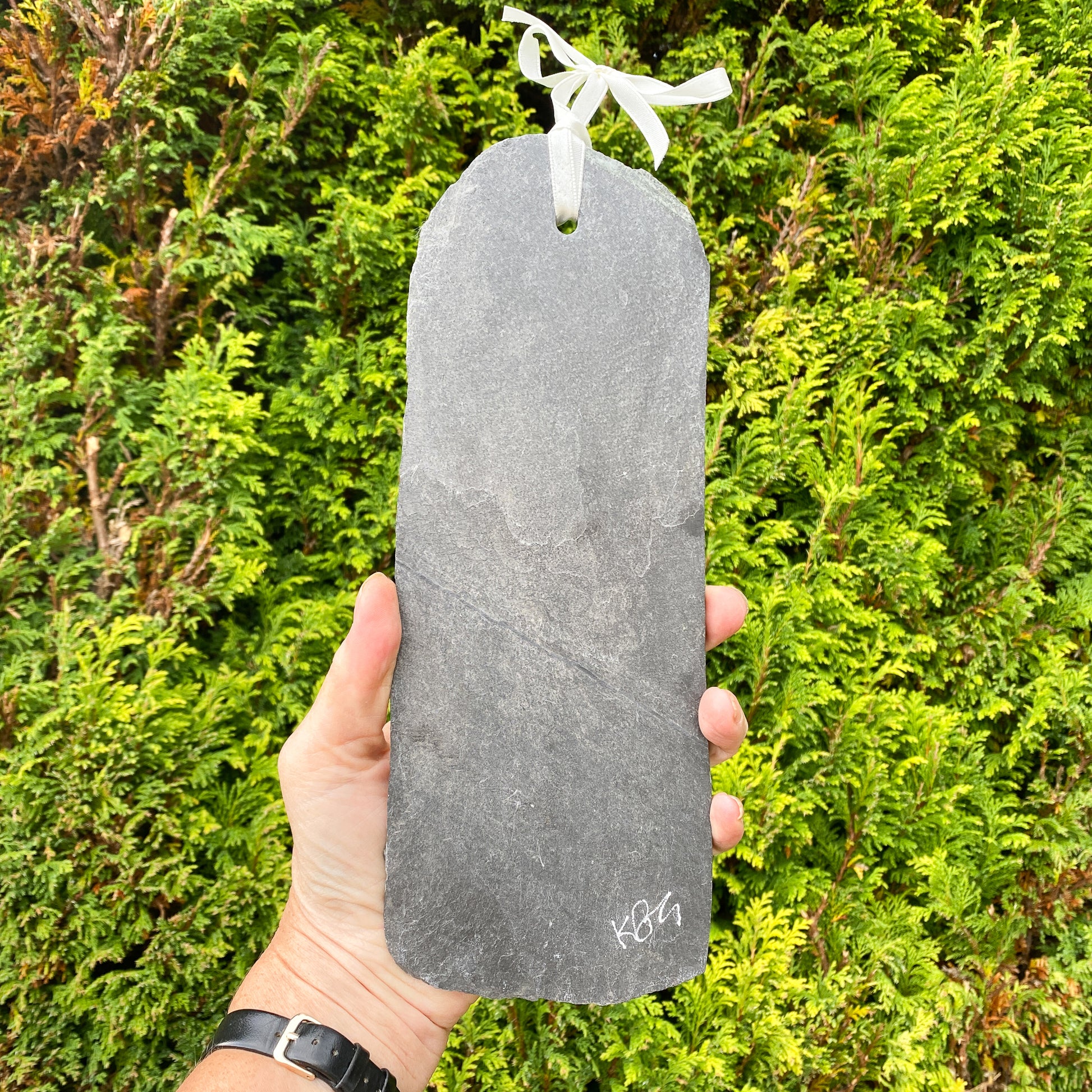 Hand holding a grey slate with a ribbon on it, with 'kbg' signature etched on it against a green bush background