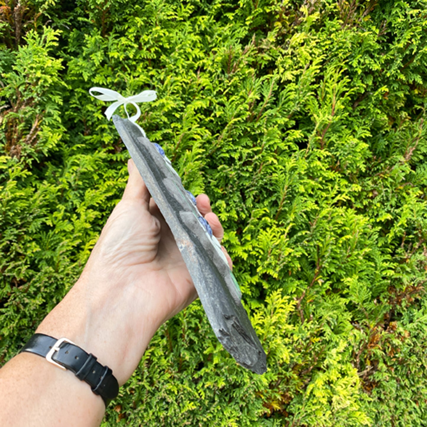 Hand holding a decorative slate mosaic with floral designs against a green bush background.  Held at an angle to show the edge of the slate.
