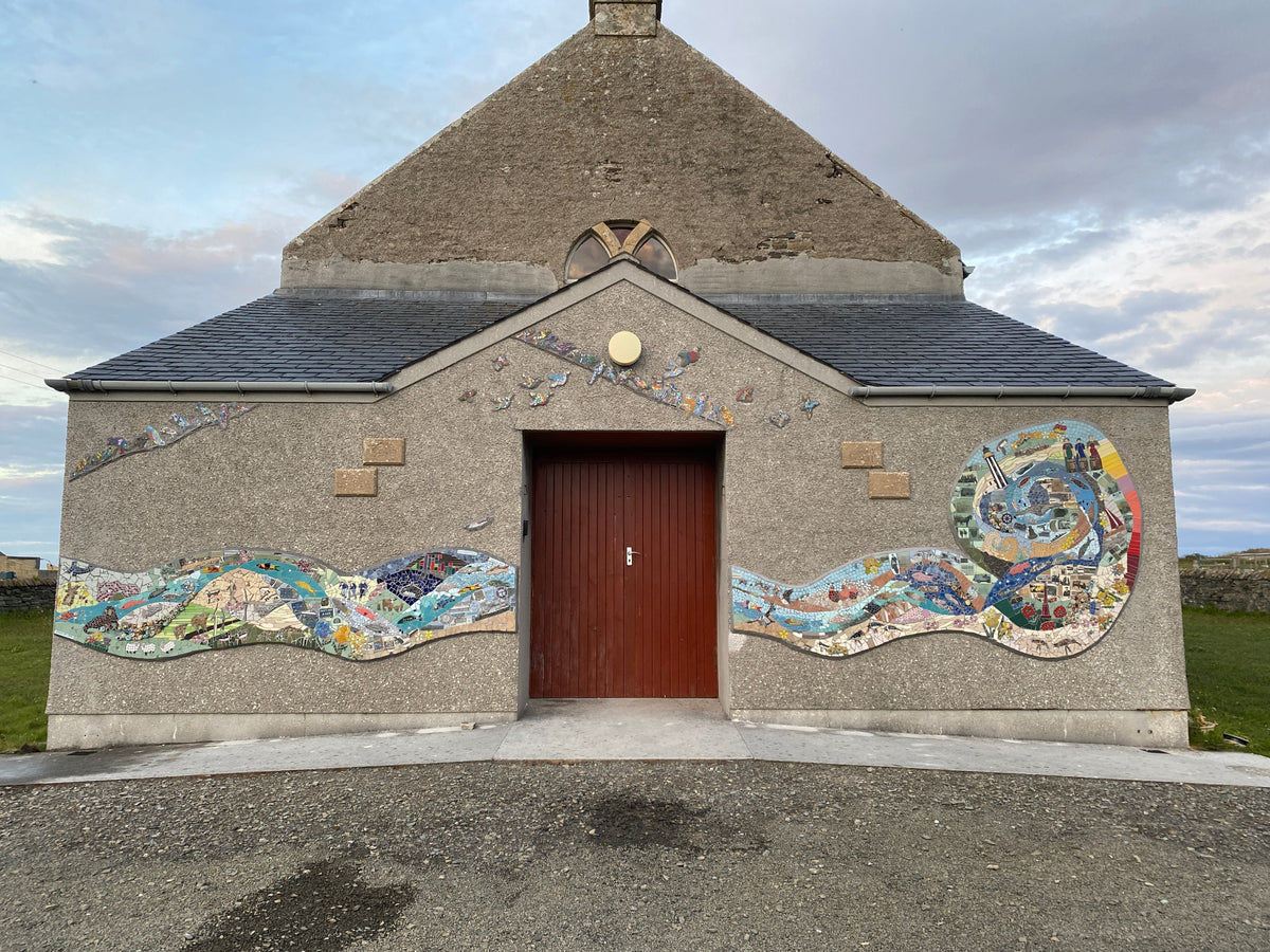 A stone chapel or small church with a dark red wooden door, decorated with colorful mosaic art on the walls and a gray slate roof, photographed outdoors under a cloudy sky.