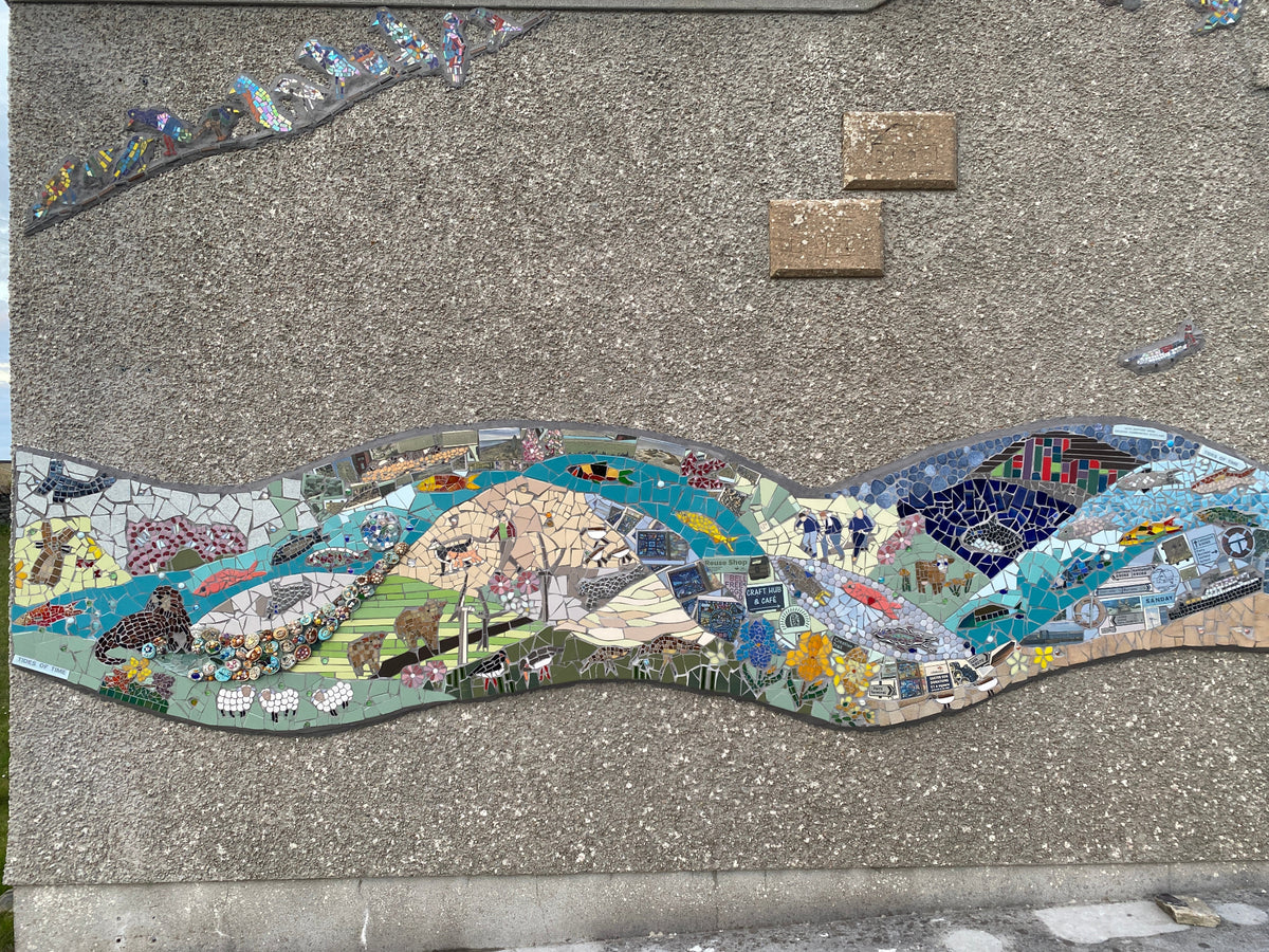A colorful mosaic wall art piece depicting a winding river landscape with boats, trees, houses, and natural scenes, mounted on a textured concrete wall.