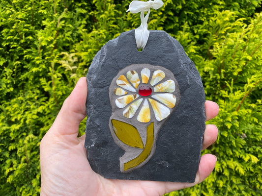 A handmade floral ornament mosaic on a dark slate stone, featuring a white and yellow flower with a red glass center, hanging from a white ribbon against a background green foliage.