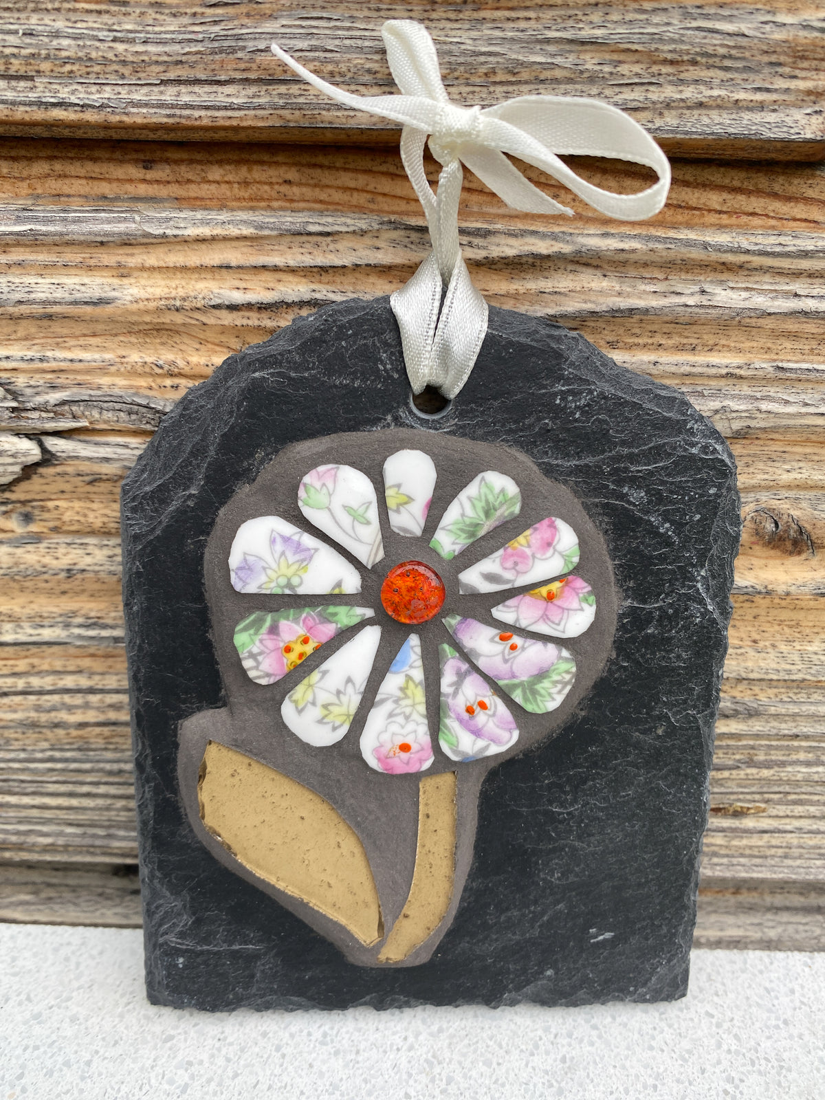 A decorative hanging ornament shaped like a flower with a dark slate backing, floral petals in pastel colors, a tan leaf shape, and a white ribbon tied at the top, shown against a rustic wood background.