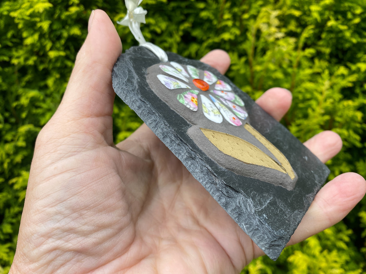 A hand holding a small rectangular slate ornament with a painted floral design and a white hanging ribbon, set against a blurred green hedge background.