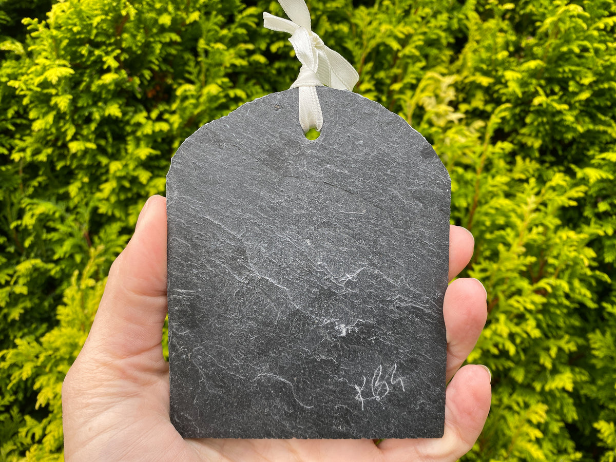 A hand holding a dark gray slate tag with a white ribbon loop, set against a background of green foliage.