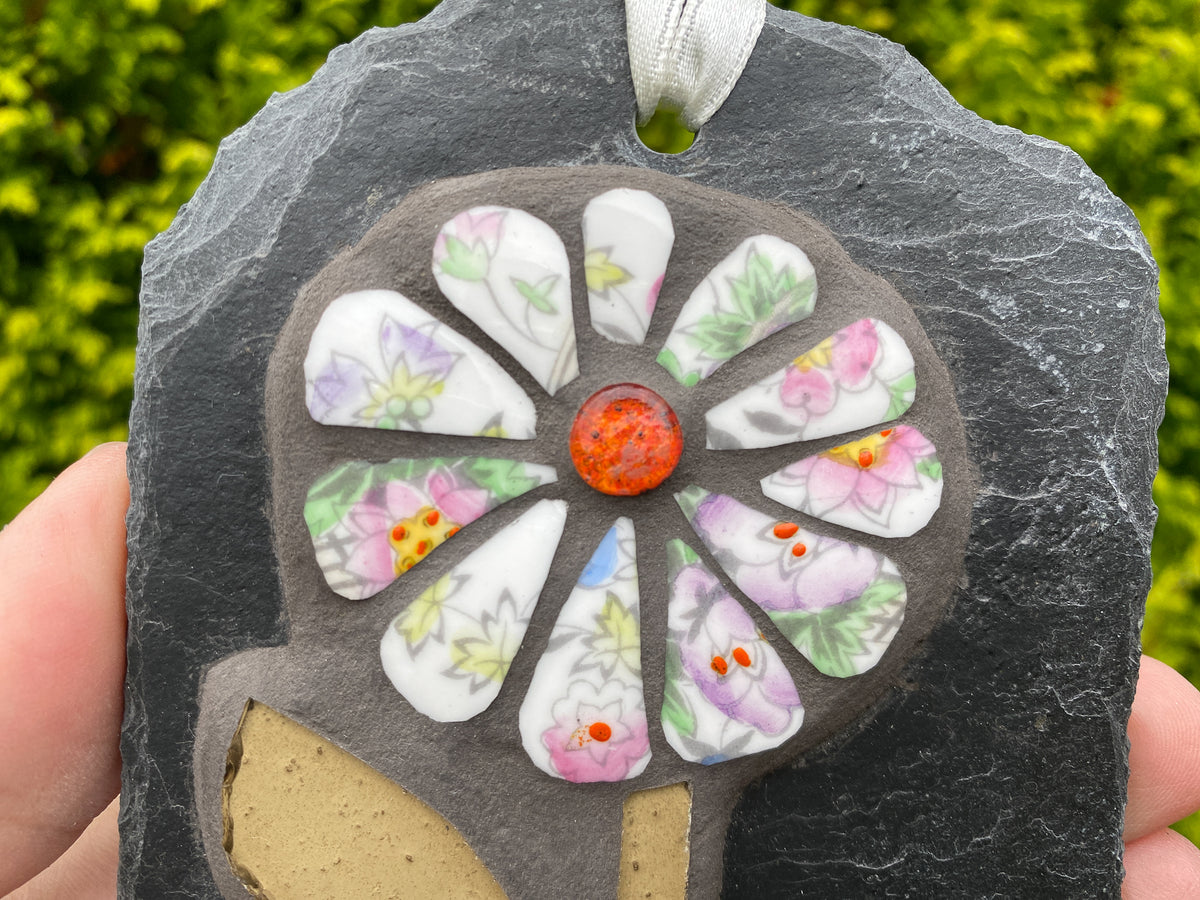 A handmade hanging ornament made from a dark gray slate, featuring a central orange glass bead surrounded by white petal-shaped tiles with pink and green floral patterns, held by hands against a blurred green garden background.