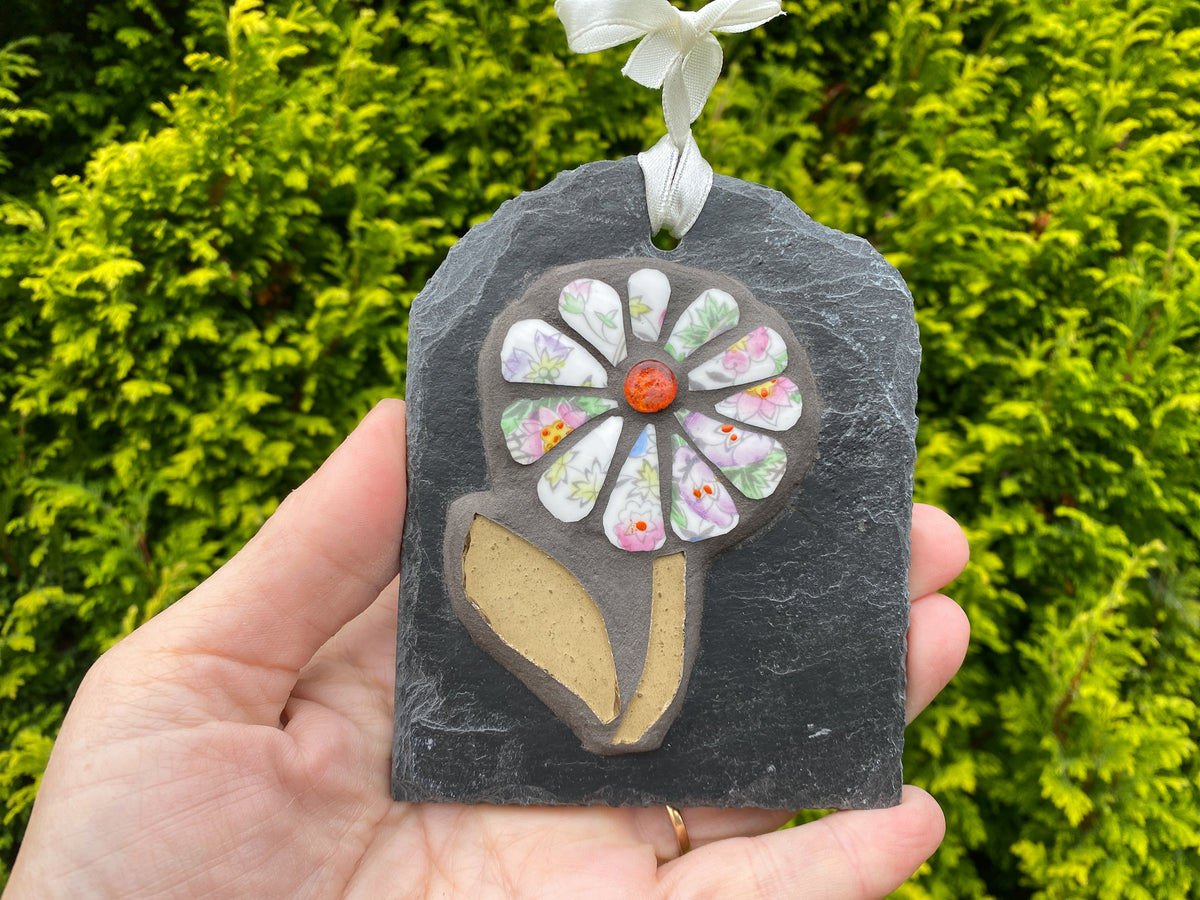 A hand holds a decorative hanging mosaic ornament shaped like a flower on a dark slate plaque, with a white ribbon loop at the top. The flower has pastel patterned petals and a small orange center, set against a background of green foliage.