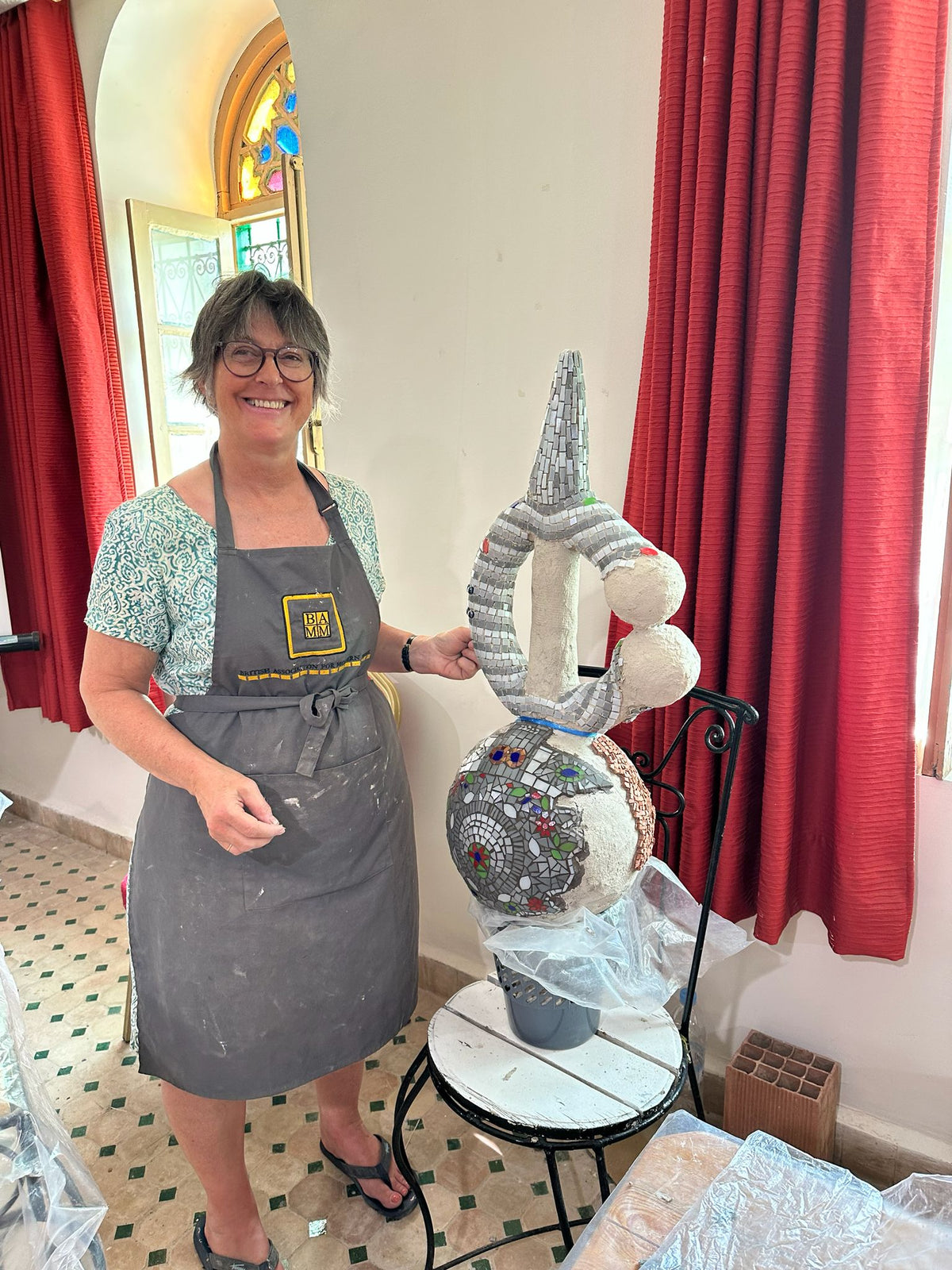 A smiling woman wearing a gray apron stands indoors next to a large, ornate sculpture with a tall pointed top and a round patterned base, set on a black metal table in front of red curtains.