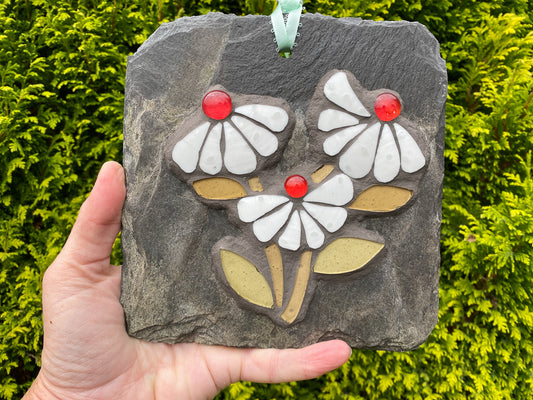A hand holds a square slate garden ornament featuring three white daisy-like flowers with red centers, white petals, and gold-toned leaves on a dark gray stone background, with green foliage behind it.