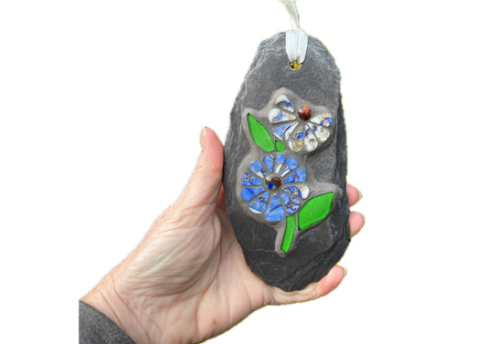 two cornflower blue and white flowers with bright green leaves in mosaic on a grey slate, hand held, with a white background