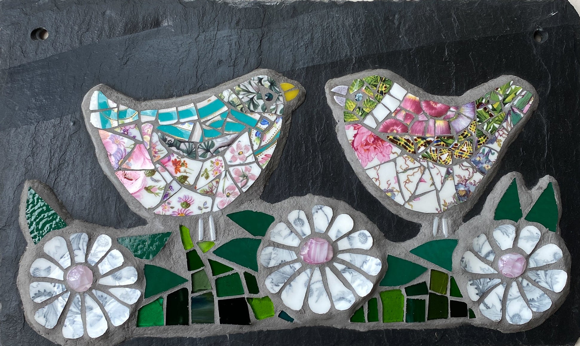 A rectangular black slate wall plaque featuring mosaic bird and flower artwork. The design shows two birds and three white flowers with green and pink mosaic details on a dark slate background, photographed against a light neutral surface.