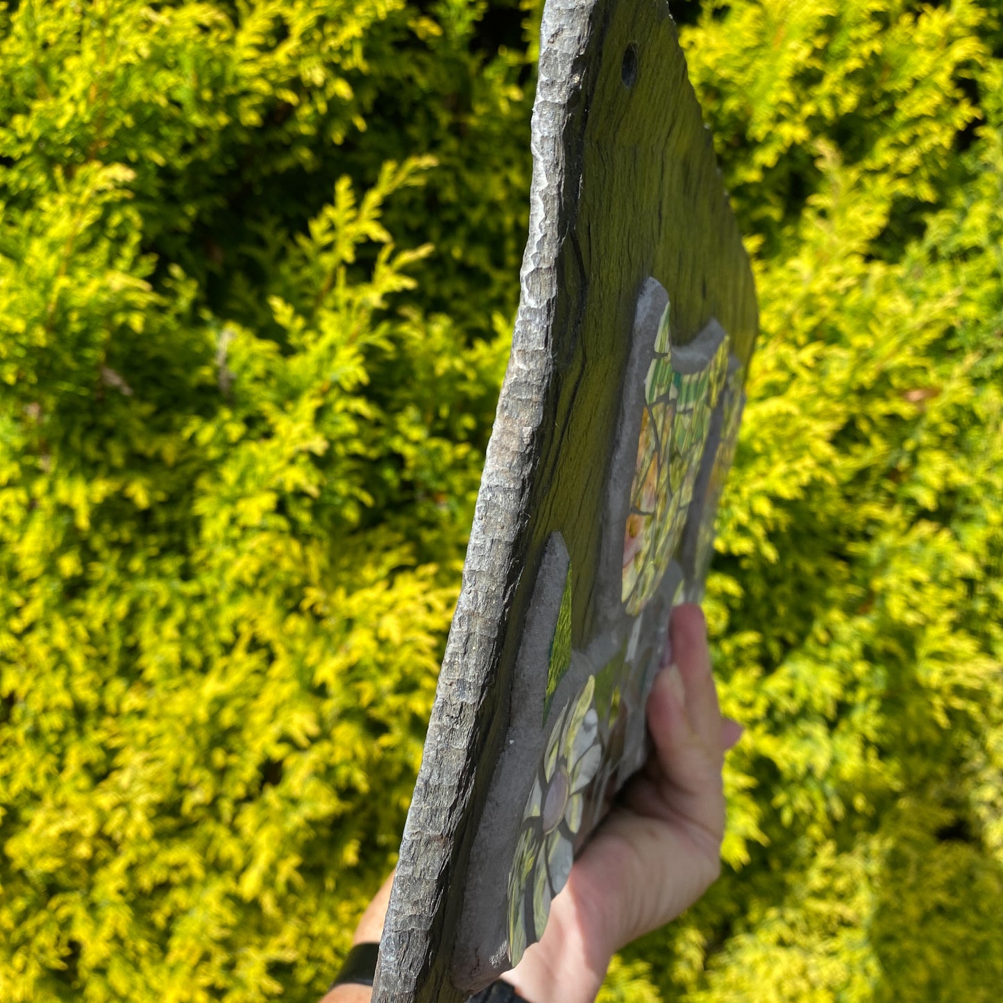 A hand holding a rectangular slate plaque with a rustic gray edge and a colorful mosaic design featuring green, yellow, and orange elements, set against a background of bright green foliage.