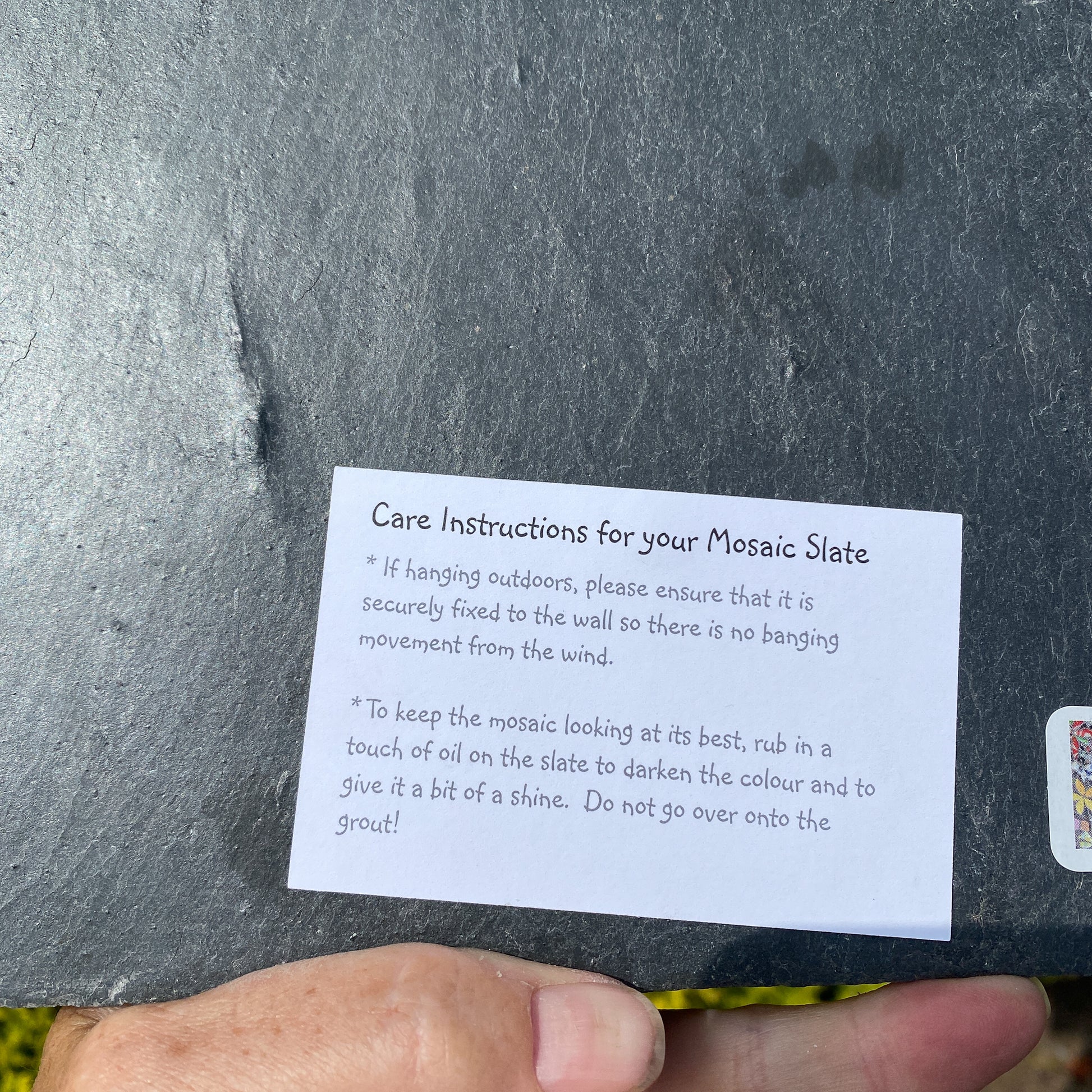 A close-up of a hand holding a piece of slate next to a small white instruction card that reads 'Care Instructions for your Mosaic Slate' with mounting and shine instructions.