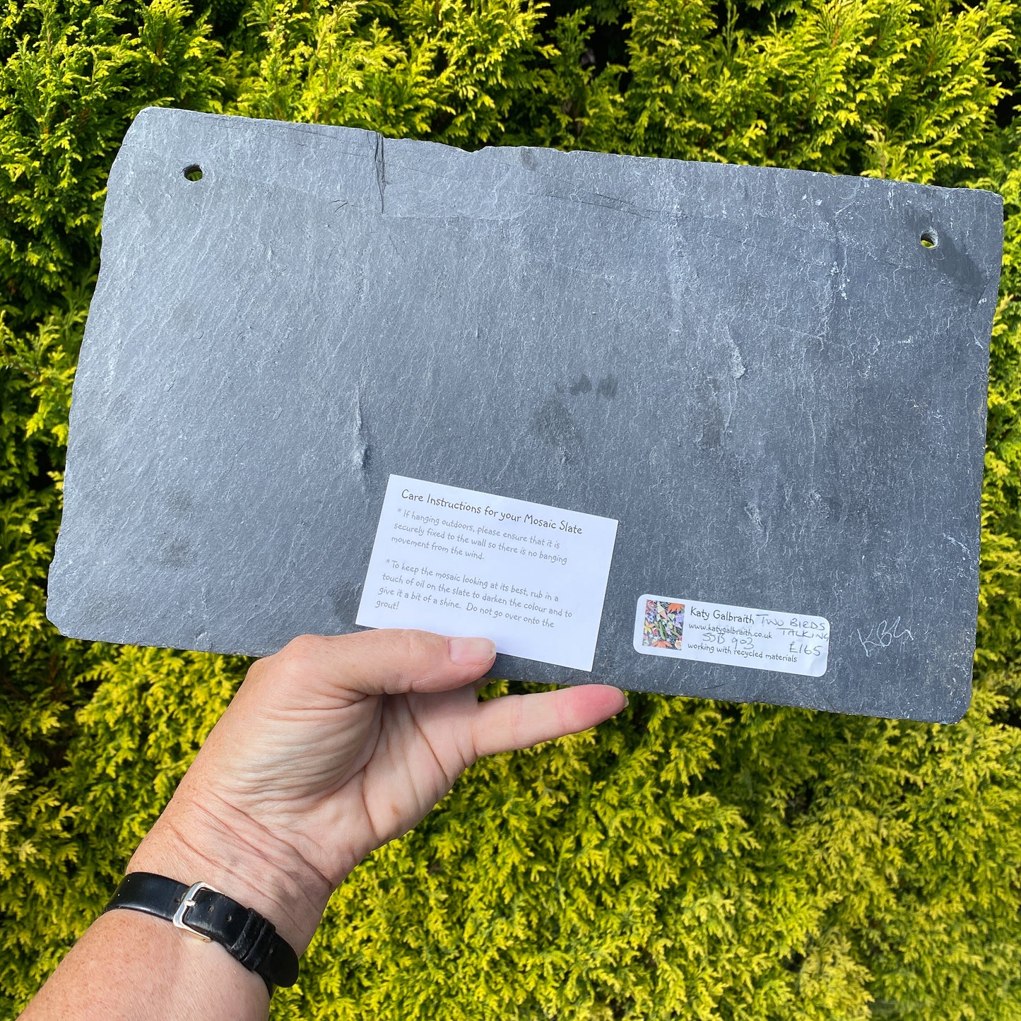 A slate plant marker or plaque being held by a hand against a background of bright green foliage. The slate has pre-drilled holes in the corners, a small white instruction card, and a small brand sticker on the bottom.