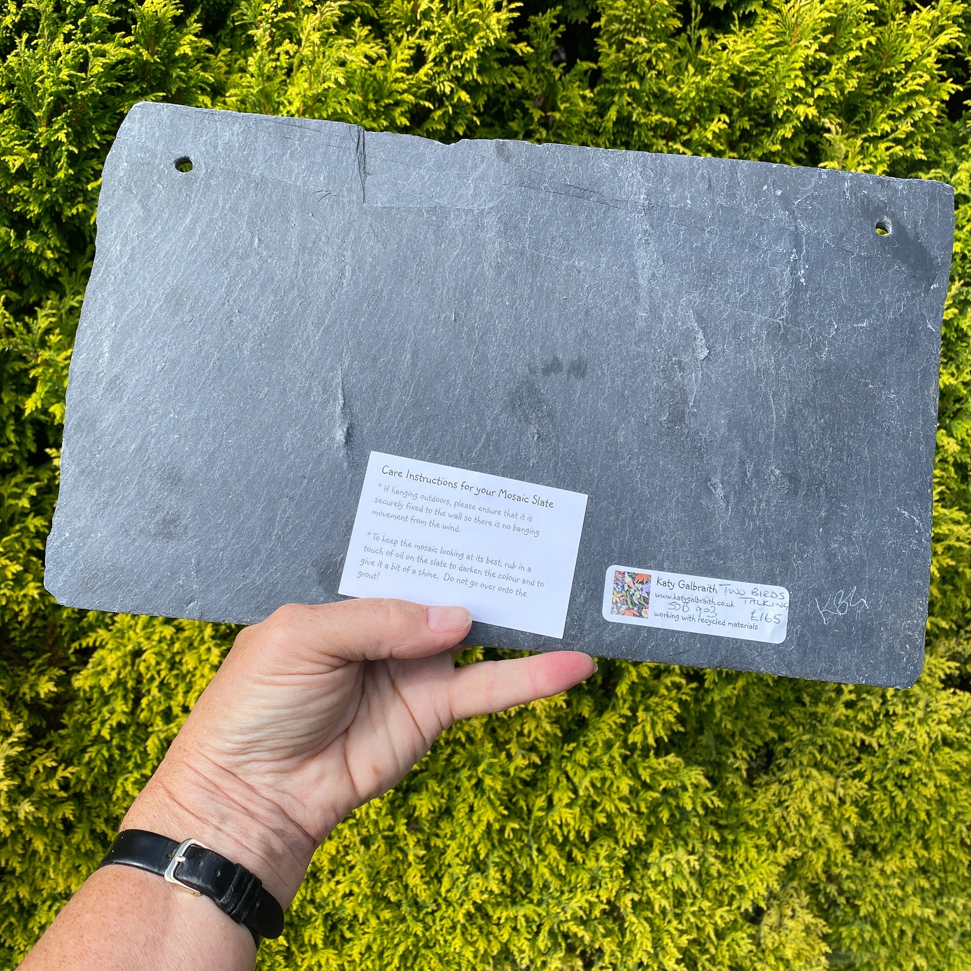 A slate plant marker or plaque being held by a hand against a background of bright green foliage. The slate has pre-drilled holes in the corners, a small white instruction card, and a small brand sticker on the bottom.