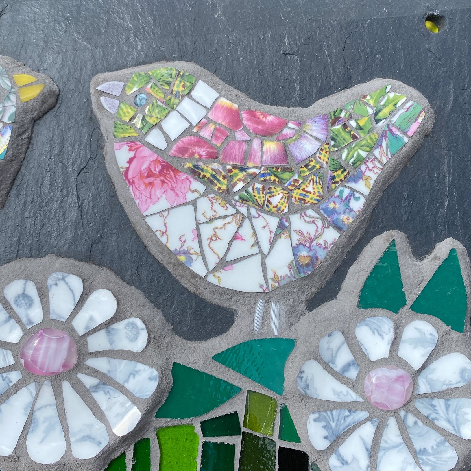 A close-up of a handmade mosaic garden stone featuring birds and flowers. The stone is made of dark charcoal slate with mosaic tiles in pink, green, white, and yellow, set against a blurred background of green foliage.
