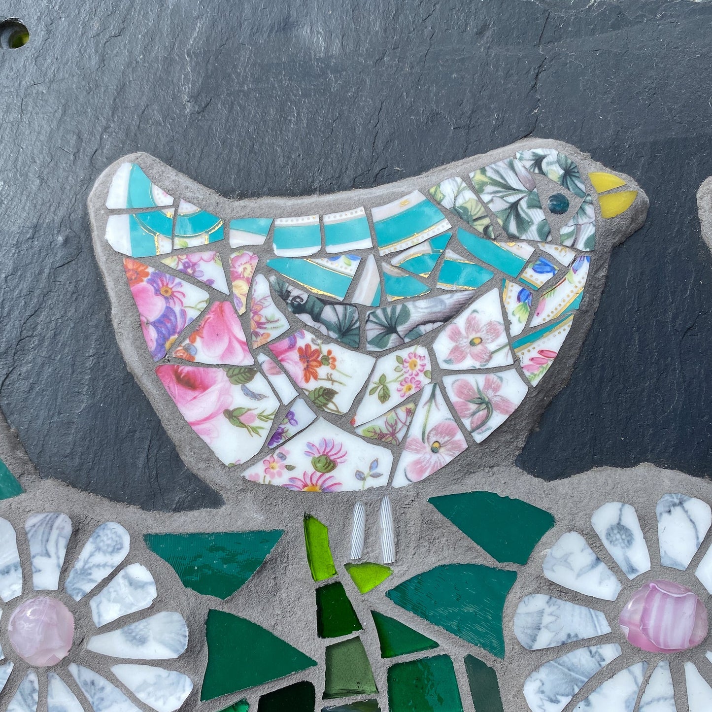 A close-up of a mosaic slate piece featuring a bird and flowers on a dark slate background.