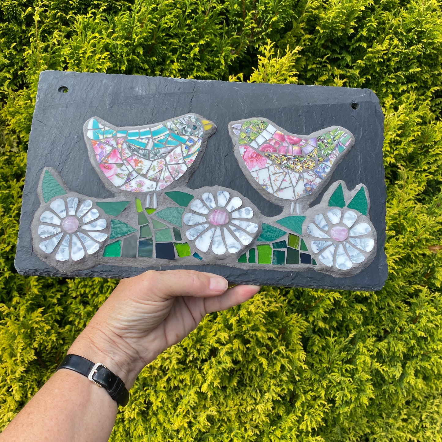A hand holds a rectangular dark slate mosaic plaque featuring two stylized birds and three white flowers with colorful mosaic details, set against a background of green conifer bushes.