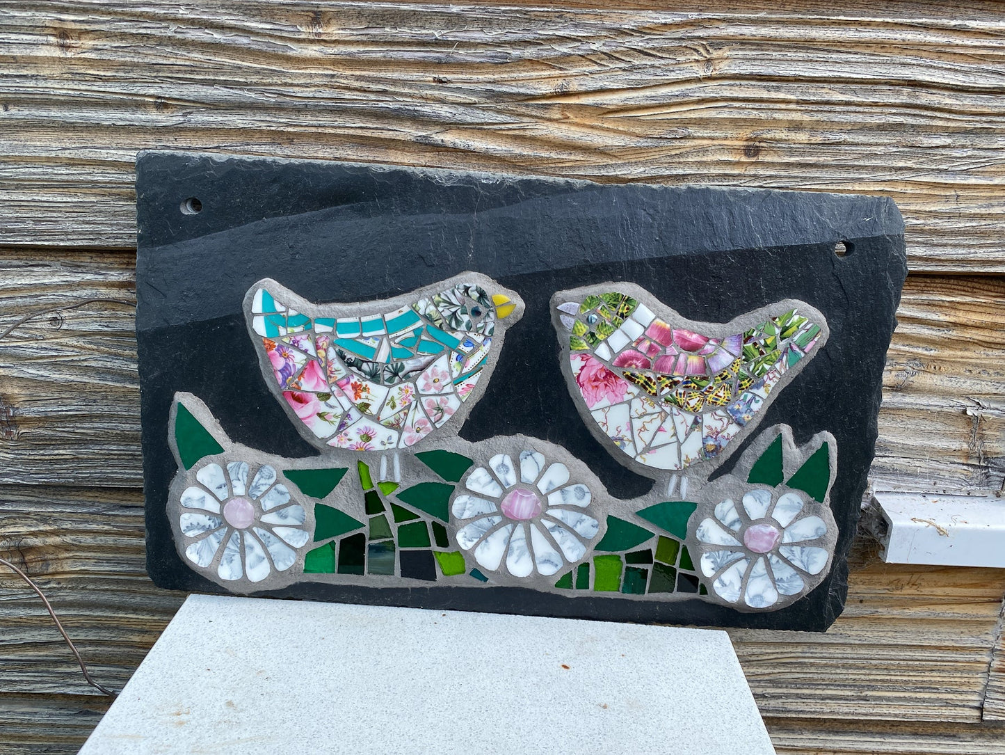 A rectangular black slate mosaic garden plaque featuring two birds and three flowers, displayed outdoors on a white stone pedestal against a weathered wooden wall.